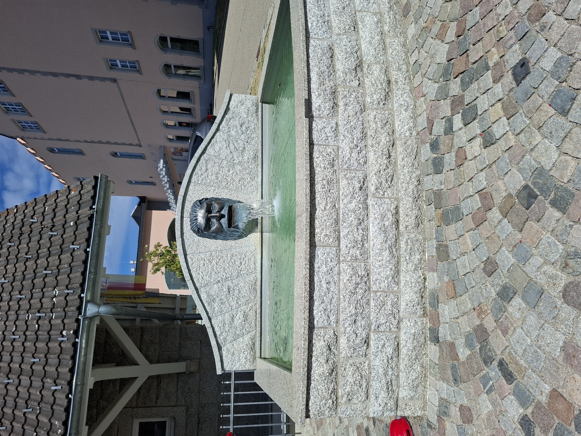 This is an eye-level, medium shot of a stone fountain with a sculpted face at its center, located in the city of Grafenhausen, Germany. The fountain has a rectangular basin filled with green water, with water spouting from the mouth of the sculpted face. The basin is constructed from light grey, rough-hewn stone blocks, and the carved face is made of a darker, metallic material, possibly bronze. Behind the fountain is a curved, granite-like wall with the same sculpted face. To the left of the fountain is a metal fence. The foreground of the image features a cobblestone pathway composed of variously colored, diamond-shaped stones. In the background, a building with a red-tiled roof and white structural elements is visible on the left, and a large, light pink building with multiple windows and arched entrances is on the right. The sky is blue with scattered white clouds. A small red object, possibly a ball, rests on the ground near the bottom left of the image.