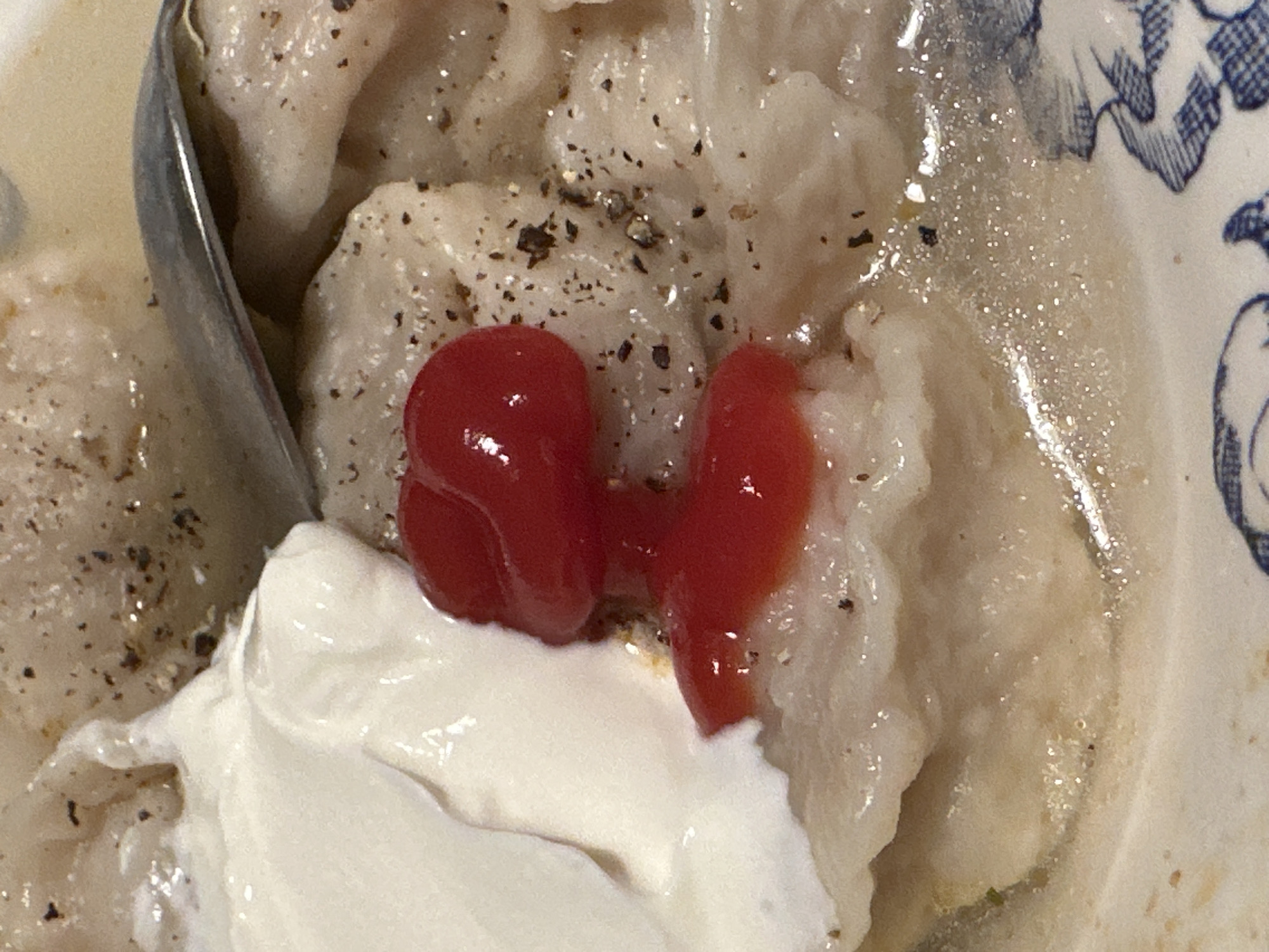 A close-up shot reveals a bowl of pierogi, garnished with black pepper, a dollop of sour cream, and two small portions of a red sauce, possibly ketchup. A spoon is partially visible on the left side of the frame. The bowl has a white base with a blue floral pattern. The exact location and city are not discernable from the image.