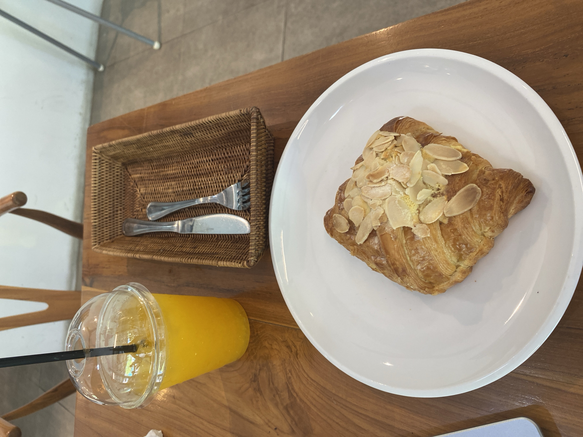A top-down view shows a wooden table arrangement with a croissant, orange juice, and cutlery. The croissant, topped with sliced almonds, sits on a white plate. Adjacent to it is a basket holding a fork and knife. A plastic cup of orange juice with a black straw is positioned near the basket. The scene appears to be set in a cafe or restaurant in North Kuta, Indonesia.