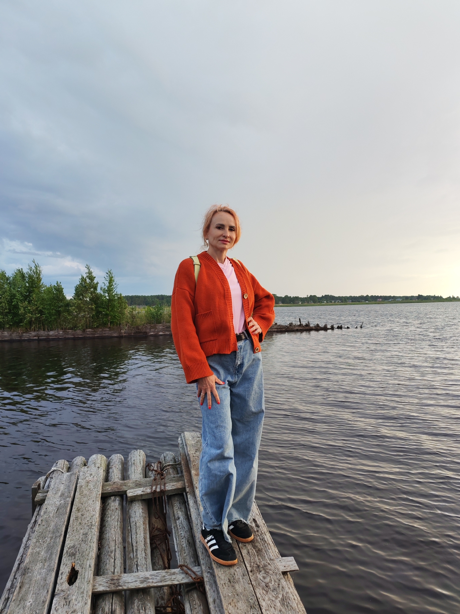 A medium shot captures a woman with blonde hair standing on a wooden pier over a body of water. She is wearing an orange cardigan over a pink t-shirt and blue jeans, and black and white sneakers. The woman has one hand on her hip and is looking at the camera. The pier is made of weathered wood planks and appears to be old and somewhat dilapidated. In the background, there is a line of trees and vegetation along the water's edge, and a hazy sky with clouds. The water is calm with ripples. The overall setting suggests a natural, possibly rural, environment. The location is identified as Shalsky, Russia, implying it is a lakeside or riverside scene within that region.