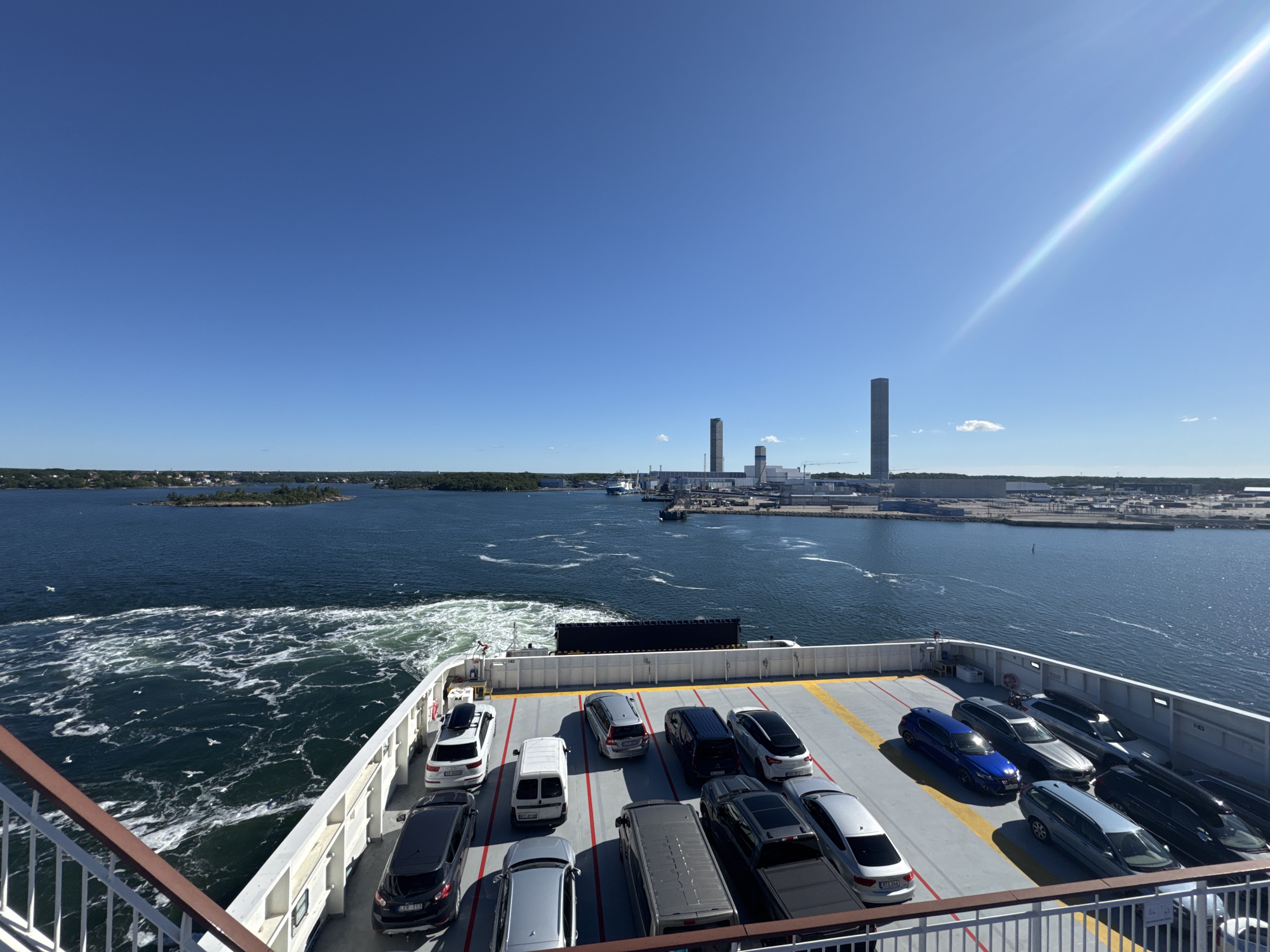 The image shows a ferry deck laden with cars, sailing on a bright, sunny day. The sky is a clear, deep blue with a prominent sun flare streaking across the upper right quadrant. In the distance, the city of Karlskrona, Sweden, is visible, characterized by industrial structures including tall, slender towers and lower buildings lining the waterfront. The water is a dark blue, with the ferry creating a white, foamy wake. Several white seagulls are scattered across the water. The ferry deck is organized with rows of parked vehicles, including SUVs, vans, and sedans. The railing of the ferry is visible in the foreground. The overall scene depicts a journey with industrial coastal scenery.