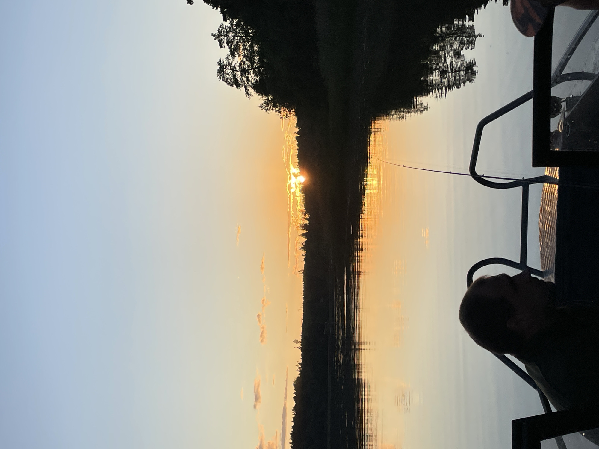 The image depicts a serene sunset over a body of water, likely a river or lake, in Berozovo, Russia. In the foreground, a person is seated in a boat, silhouetted against the water. A fishing rod is visible, planted in the boat, suggesting a fishing activity. The sunset casts a warm orange glow across the sky and reflects vividly on the water's surface, creating a mirror-like effect. The background features a dense line of trees, forming a dark silhouette against the vibrant sky. The overall atmosphere is tranquil and picturesque.