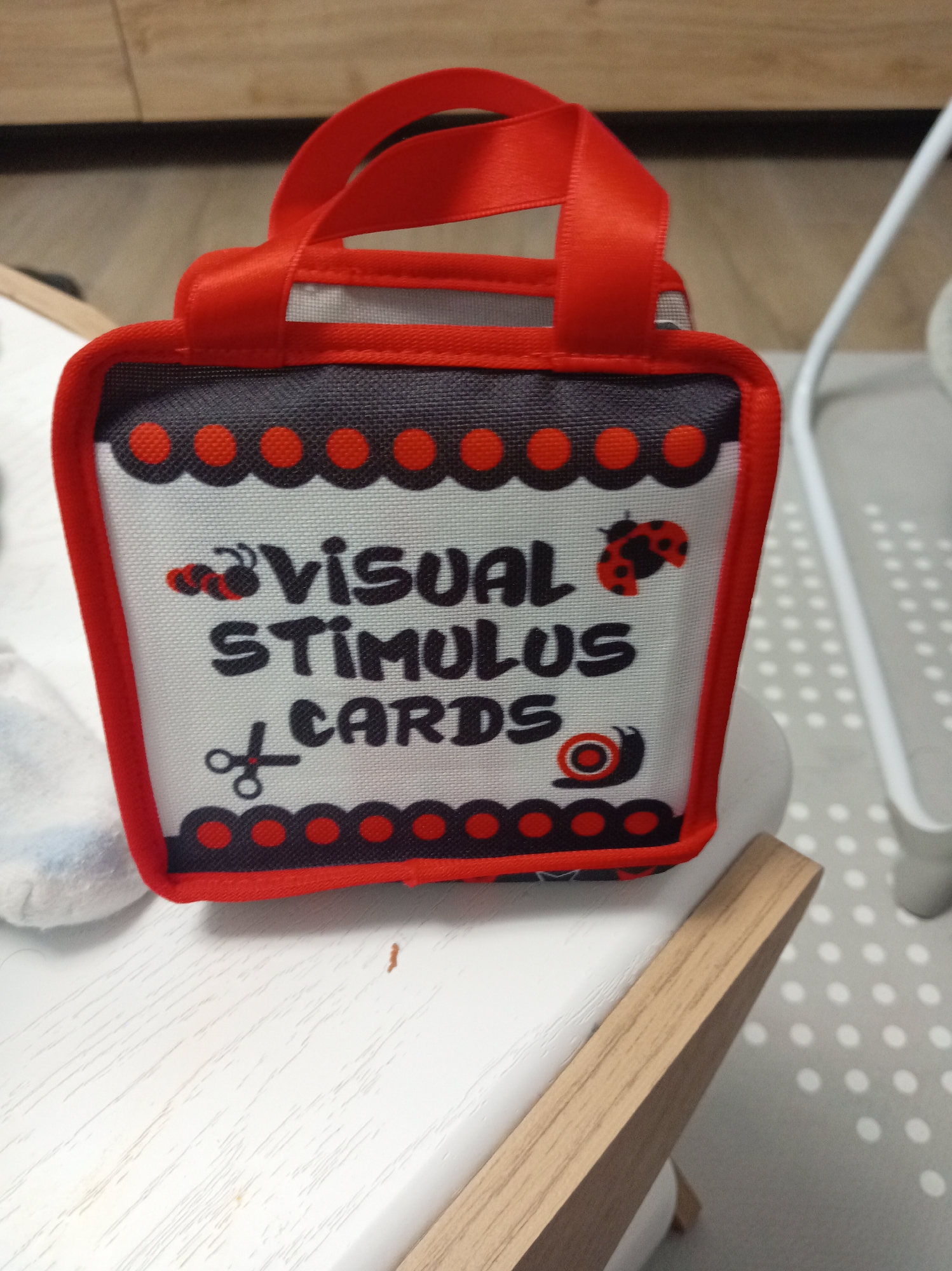 A close-up, slightly angled overhead shot shows a small, red-handled fabric bag with "VISUAL STIMULUS CARDS" printed on it in black lettering. The bag has a black and red polka-dot trim along the top and bottom edges and features small ladybug and snail illustrations. It is placed on a white, wooden surface that appears to be part of a chair or table, with a light-colored, textured mat and a white object with a light-colored cord in the background. The overall scene is indoors, with a blurred wooden surface in the far background. The image is set in Unknown, Unknown.