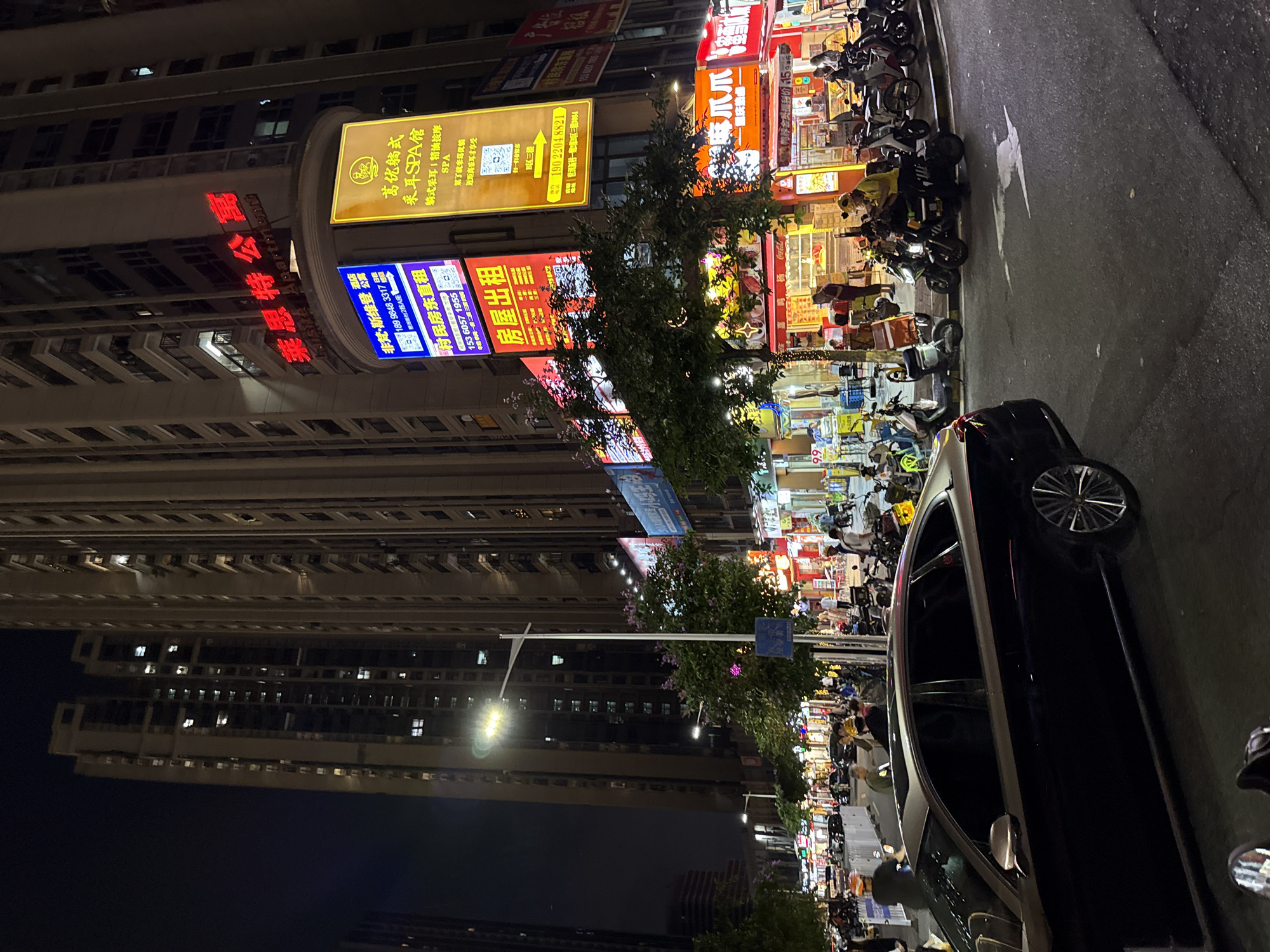 This image depicts an urban scene in the Haizhu District of Guangzhou, China, at night. High-rise buildings dominate the left side of the frame, displaying various illuminated signs in Chinese characters, including "莱思特公寓 Lisite Apartment," "非梵·斯维登," "村民房东直租," "房屋出租," and "葛优躺式 采耳SPA馆."
On the right side, a row of storefronts with bright, colorful signs indicates commercial activity. A street lined with parked motorcycles is visible. The foreground is occupied by a dark car, its details partially obscured. People are present in the scene, interacting with the businesses. A street light shines in the middle distance. The overall environment suggests a bustling commercial area at night.