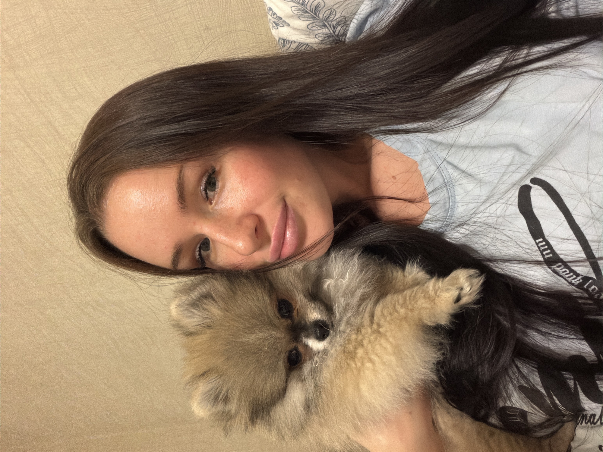A heartwarming, close-up indoor portrait captures a young woman and a fluffy Pomeranian dog. The woman, with long, straight dark brown hair, is seen from the chest up, her head gently tilted and resting against the dog's fur. She has a soft, serene smile and is looking directly at the camera. Her complexion is clear, with a natural, rosy blush on her cheeks. She wears a light blue top, possibly a t-shirt, with some partially visible black lettering on the right side, which appears to include fragmented words like "mmm pop punk lo" and "onal."
The small, light brown and cream-colored Pomeranian dog is held in her arms, its face prominently featured in the lower-center of the frame, also looking towards the camera. Its fur is soft and voluminous, and it has dark, expressive eyes and a small black nose. One of its paws is visible, nestled in the woman's hair.
The background consists of a plain, light beige wall with a subtle textured pattern. Behind the woman's head, a light-colored pillow with dark, intricate leaf or floral patterns is visible, suggesting a cozy, domestic setting like a bedroom or living room. The image conveys a tender moment of companionship and affection between the woman and her pet. The lighting is soft and even, highlighting their features. No specific time of day or outdoor weather conditions can be determined from this indoor scene.