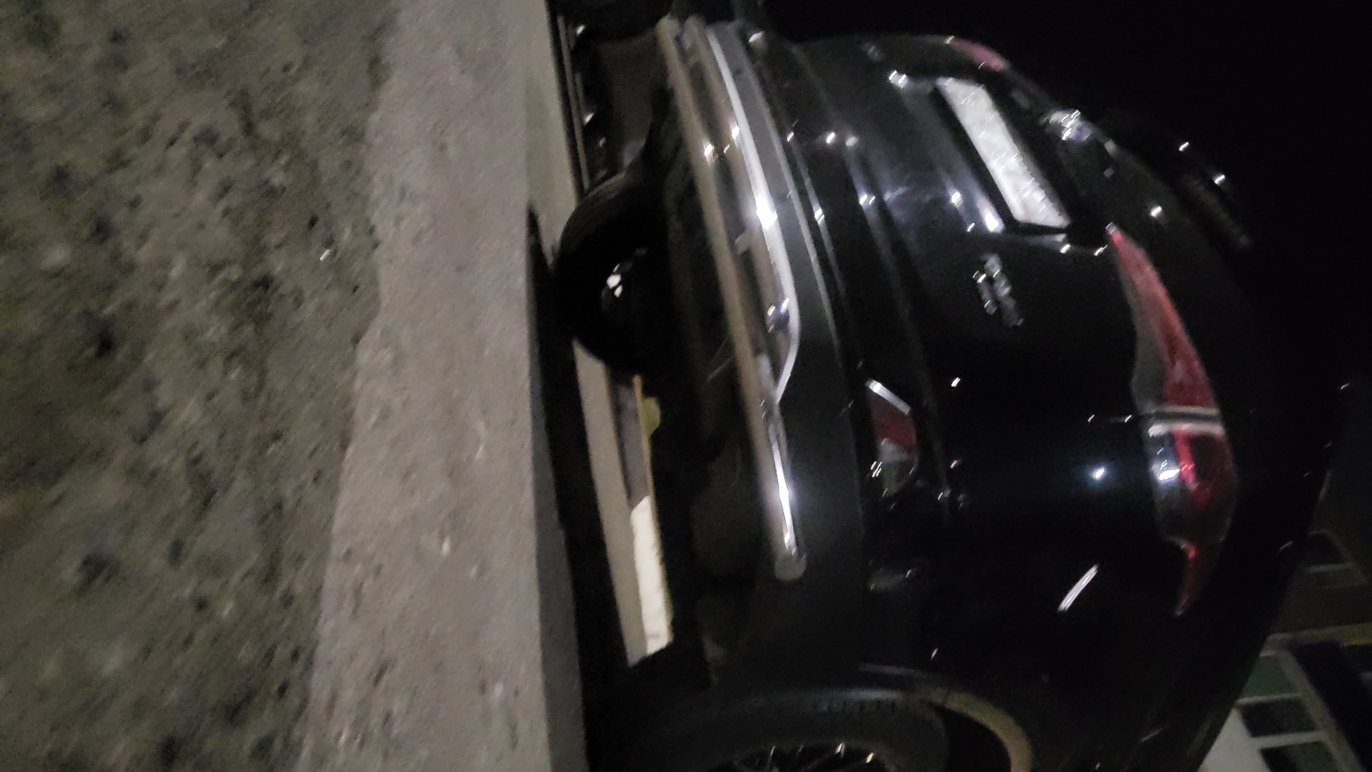 A low-angle, slightly tilted photograph shows the rear quarter of a black car parked on a paved surface. The car features chrome accents and a distinctive rear taillight design. The location is not evident from the provided image alone.