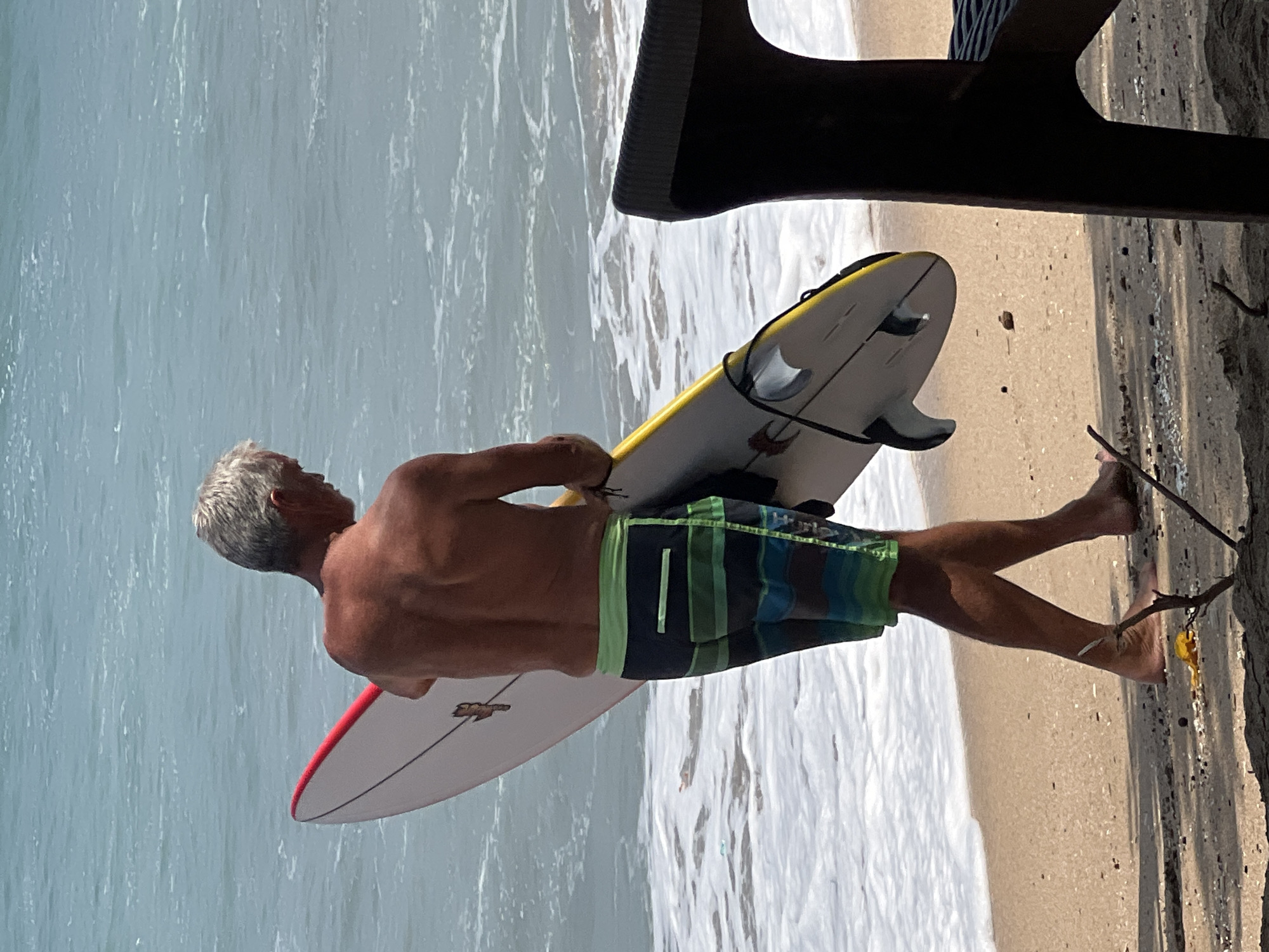 An adult male with grey hair, wearing multi-colored striped swim trunks, stands on a sandy beach near the water's edge, holding a white surfboard with a red stripe along the edge. The surfboard has black fins visible. The man is viewed from behind, and he is looking down. The ocean behind him is calm with small waves. In the foreground, a portion of a dark brown chair is visible on the right side of the frame. The scene appears to be in Kuta, Indonesia.