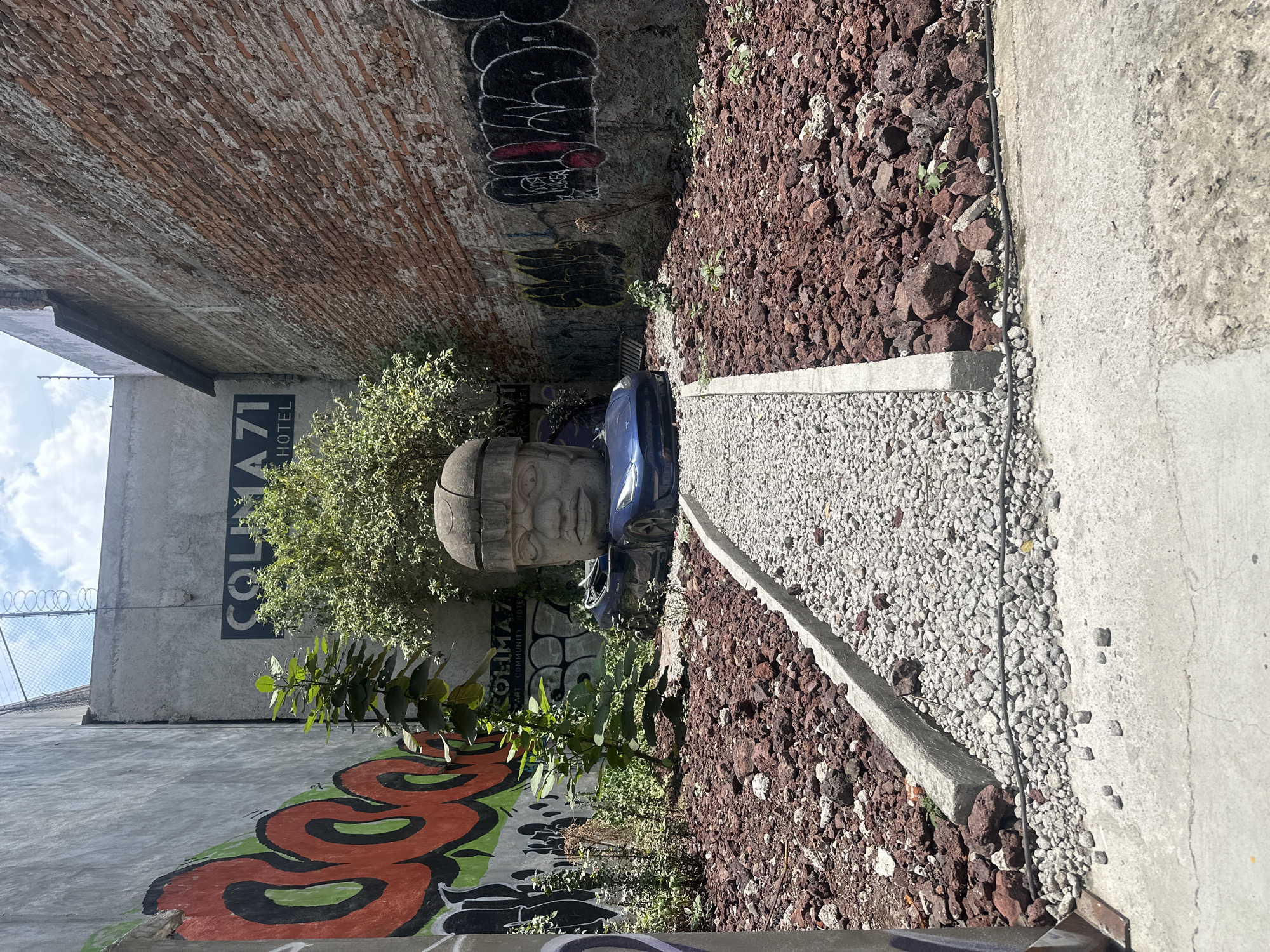 The image shows an outdoor scene in Mexico City, Mexico. A grey Olmec head sculpture sits on a blue sedan in a narrow space between buildings. The sculpture is located near the Colima71 Art Community Hotel building, and is flanked by green shrubs. The ground is covered with red volcanic rock and a narrow gravel path. Graffiti covers the surrounding brick and concrete walls.