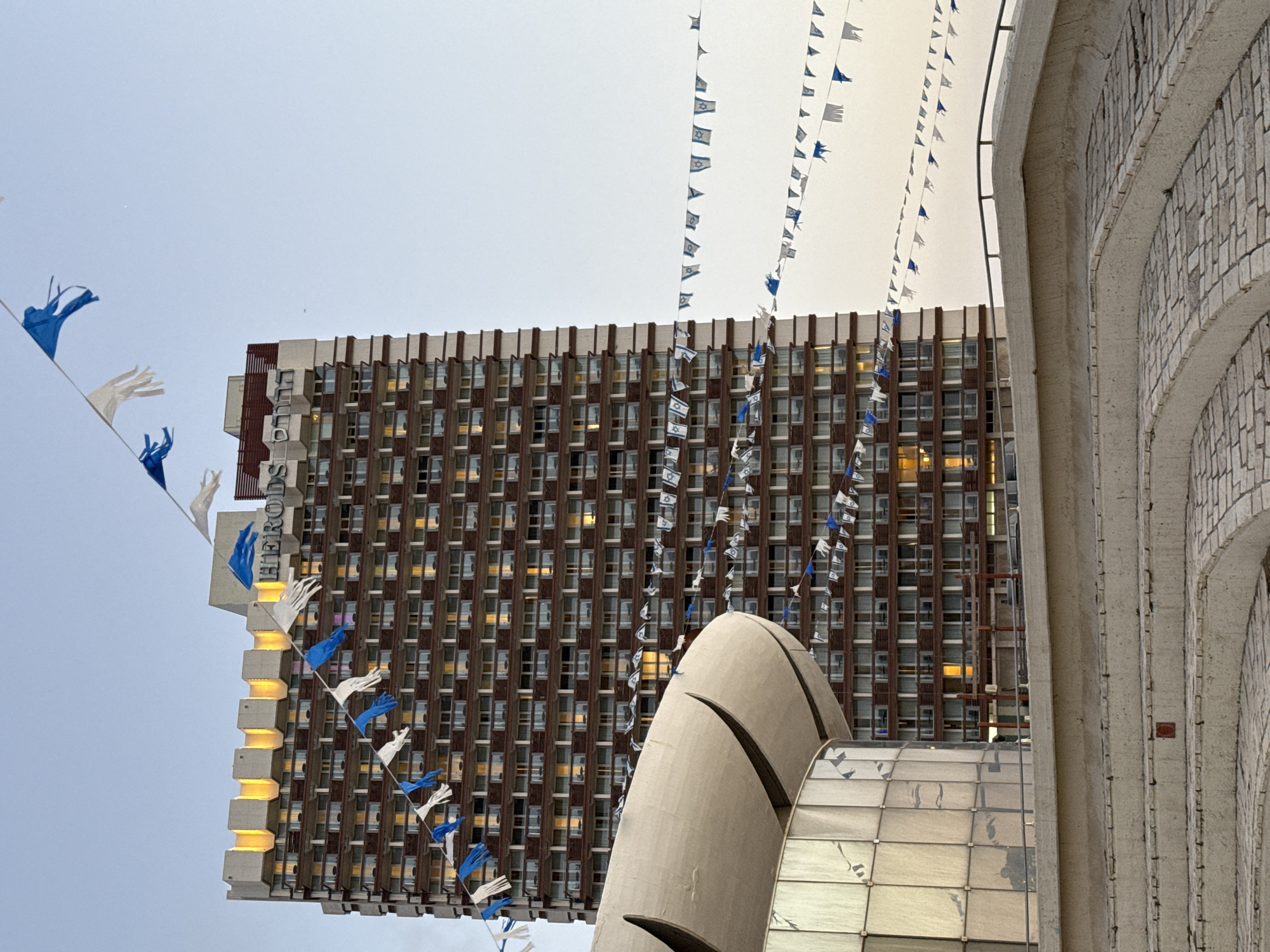 The image displays a vertical composition featuring a tall, rectangular building with several architectural elements and numerous decorative flags, set against an overcast sky in what appears to be an urban environment in Tel-Aviv, Israel.
The central subject is a multi-story building, likely a hotel or office block, characterized by a grid of small, regularly spaced windows. Many of these windows show interior lights glowing a warm yellow. The building's facade is primarily brown and grey. Along its left edge, the word "HERODS" is visible, written vertically in white capital letters on a darker background, suggesting it is the Herods Tel Aviv hotel.
Numerous strings of small flags, predominantly blue and white, crisscross the upper and central parts of the frame. Many of these flags clearly depict the Star of David, a national symbol of Israel, indicating a festive or patriotic occasion. Other flags are plain blue or white.
In the foreground, towards the bottom and right, are parts of other structures. On the far right, a large, curved concrete wall, possibly part of an arched facade, features a textured, tiled surface. Below the main building, a large, smooth, light-grey, egg-shaped or abstract sculptural element with distinct seams is partially visible. Adjacent to this, a sloped glass structure, likely a roof or skylight, reflects the ambient light.
The sky is a uniform, light grey, suggesting an overcast day. The illuminated windows in the building contrast with the soft outdoor light, implying it might be late afternoon or evening. The overall scene conveys a sense of modern urban architecture adorned for a national celebration.