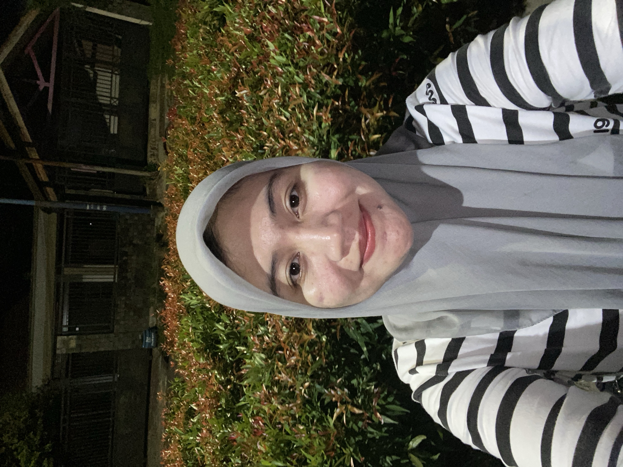 A medium shot selfie features a woman wearing a light grey hijab and a black and white striped long-sleeved shirt. She is positioned in front of a dense hedge with reddish-brown and green foliage. Behind the hedge, a dark, low-lit structure with a sloped roof and visible supports can be seen. The overall scene is dimly lit, suggesting it was taken at night. The woman's face is the primary focus, with her gaze directed towards the camera and a slight smile. The background appears to be an outdoor setting, possibly a garden or park area. The presence of artificial lighting is suggested by the illumination of the woman and the foliage, while the background structure remains largely in shadow. The scene does not provide specific landmarks that would definitively place it in Banjarbaru, Indonesia, but the foliage and architectural style are consistent with tropical or subtropical environments.