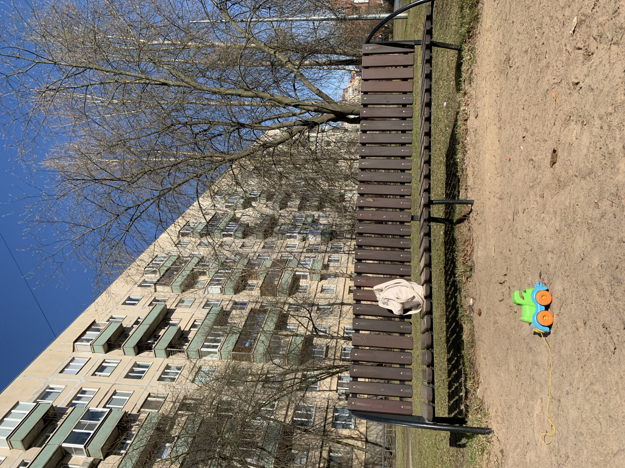 The image depicts a street scene in Saint Petersburg, Russia. A brown wooden park bench sits on a patch of grass next to a dirt area. A white tote bag rests on the bench. A colorful plastic toy truck with a yellow pull string lies on the dirt. Behind the bench is a tall apartment building with numerous balconies and windows. Bare trees stand in front of the building, and a clear blue sky is visible.