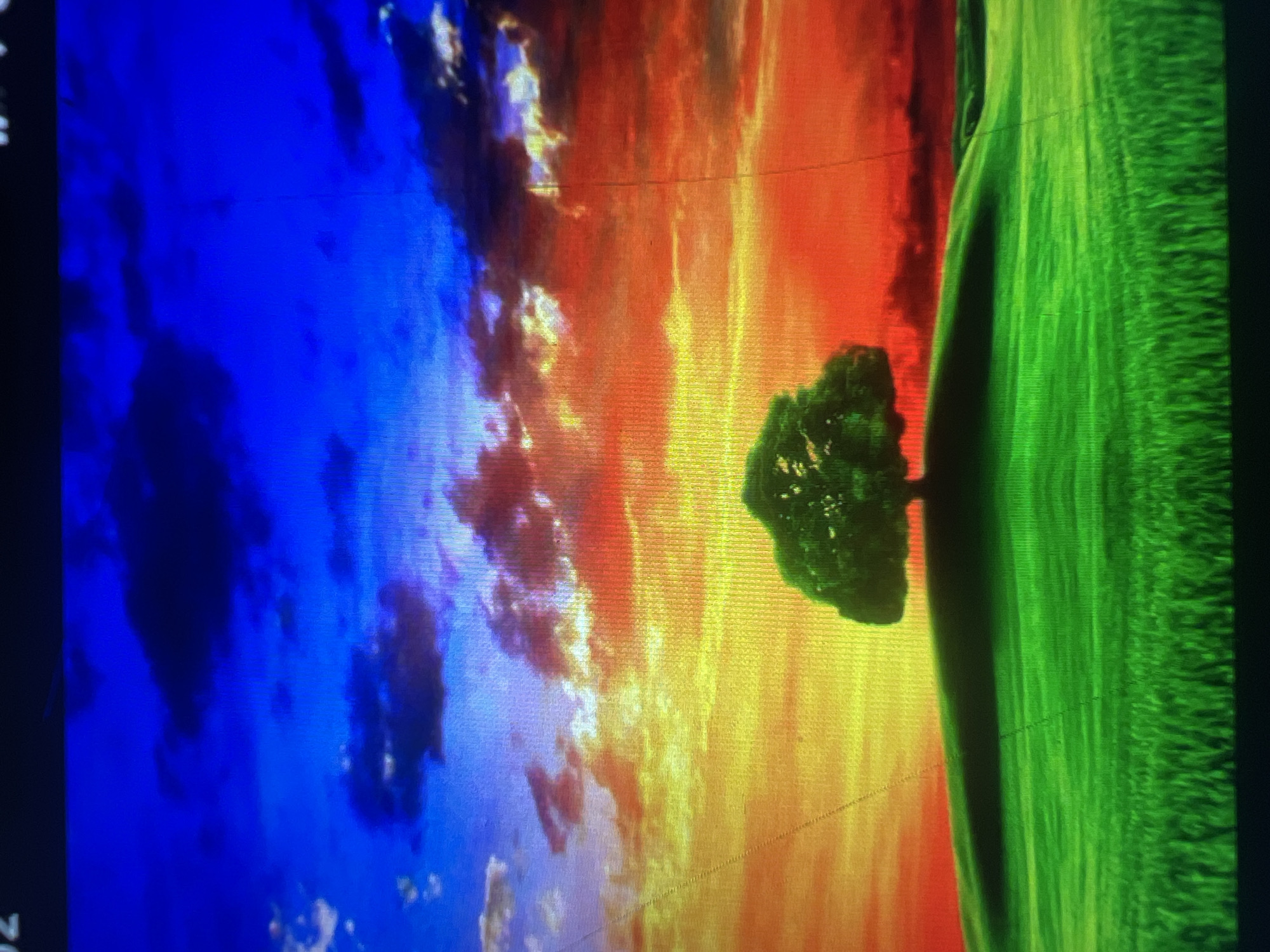 The image depicts a vibrant and stylized landscape with a lone, lush green tree silhouetted against a dramatic sunset. The sky transitions from a deep, rich blue at the top to fiery hues of orange and red near the horizon, with scattered clouds tinged with pink and yellow. Below the sky, rolling green hills create a serene foreground, with a textured representation of grass. The overall impression is one of intense color and natural beauty, likely a digital artwork or heavily edited photograph. The scene is devoid of any human presence or structures. The exact location is unknown.