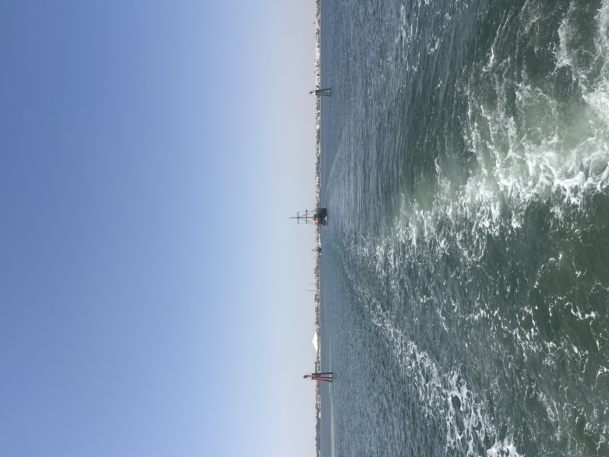 The image shows a view from a boat looking out at the sea towards a coastal city. The foreground is dominated by the foamy wake left by the boat. In the middle ground, a tall, dark sailing ship with multiple masts is visible, moving away from the viewer. To the left of the sailing ship, a red and white buoy stands in the water. To the right, a green and white buoy is also present. The background features a sprawling coastline with numerous buildings, suggesting a populated area. The sky above is a clear, bright blue, indicating a sunny day. The location is identified as the sea near Houmt Souk, Tunisia, with the coastline possibly representing the city's edge. There are no people clearly visible in the image.