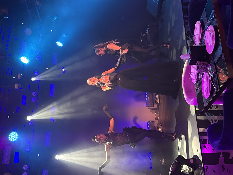 A live performance is underway on a stage within an indoor entertainment venue in Dubai, United Arab Emirates. Three female performers are illuminated by blue and white theatrical spotlights. The central figure, a vocalist, is actively singing into a microphone, wearing a black sleeveless top, wide-leg black trousers, a belt with a large buckle, long black gloves, and a multi-strand silver-colored necklace. To her left, a dancer is positioned with arms extended, dressed in a black embellished top with feather accents, black leggings, and black high-heeled boots, also wearing long black gloves. To the right, another dancer is in a similar black embellished outfit with feather details, black leggings, high-heeled boots, and long black gloves, engaged in a pose. Overhead and on the stage floor, various stage lights, including LED par cans and moving heads, emit strong beams. In the immediate foreground, part of the audience area is visible, featuring a square dark-surfaced dining table set for two with pink plates, silverware, wine glasses, and a black napkin dispenser. The chairs are upholstered in dark purple velvet. The floor directly in front of the stage exhibits a black and white geometric pattern. Ceiling-mounted security cameras or fixtures are visible above the stage area.