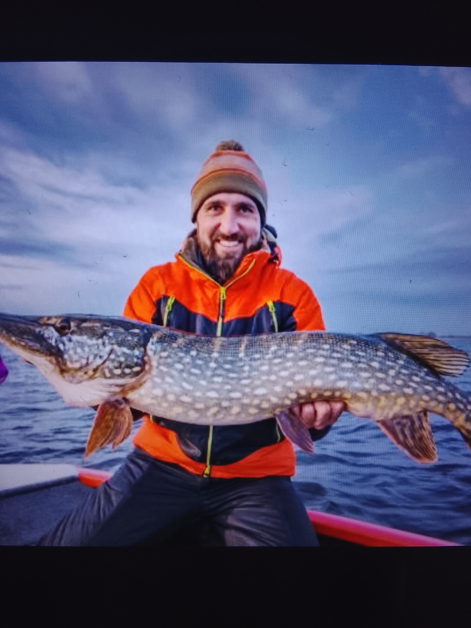 A man in an orange and black jacket and a beanie hat holds a large, spotted fish in front of him. He is smiling and standing in a boat on the water. The sky is blue with some clouds. The image does not provide any information about the city of Prisel'e, Russia, or its location.