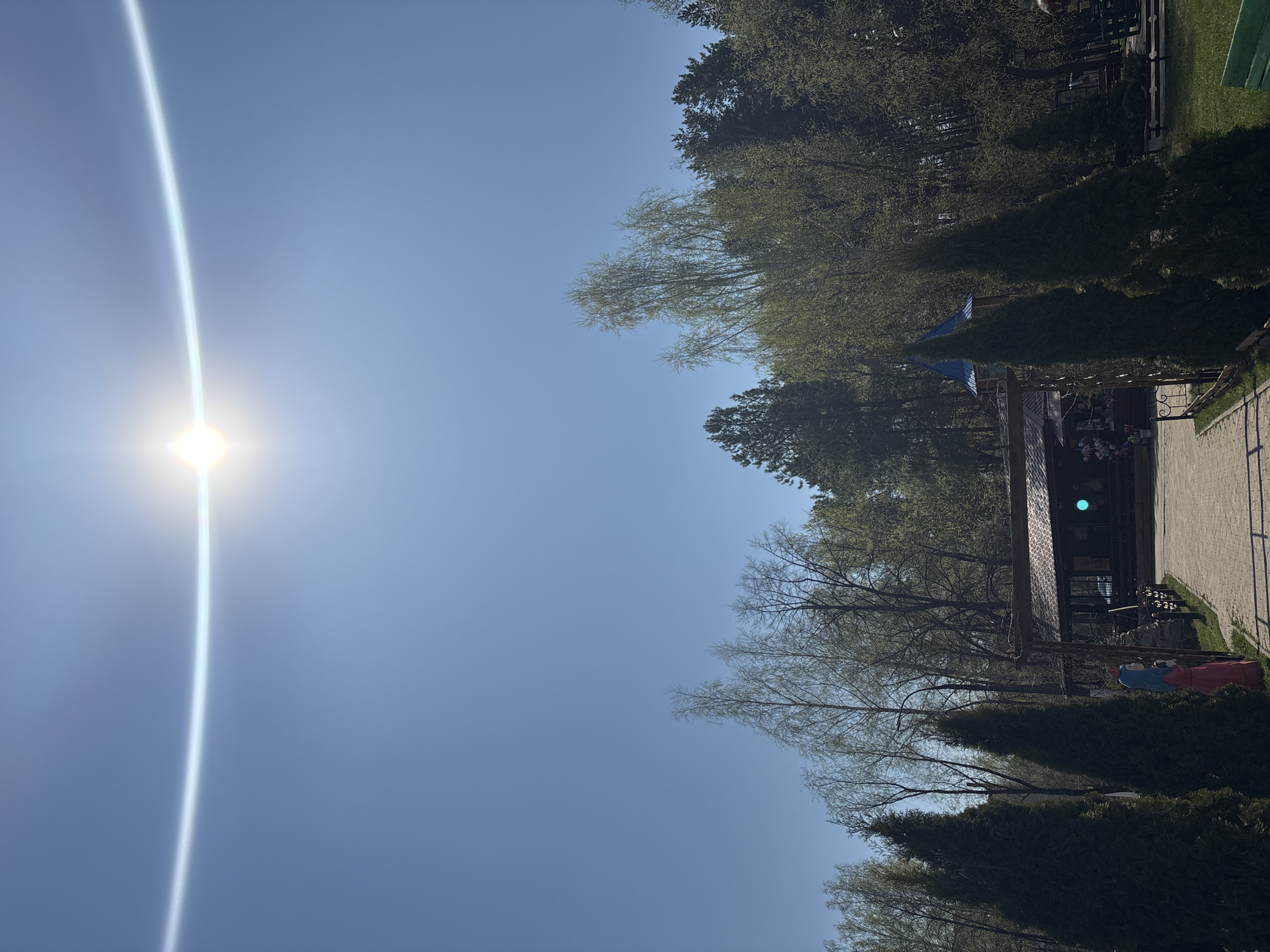 The image shows an outdoor scene in Chaikovsky, Russia, taken on a bright, sunny day with the sun prominently visible. The sky is clear blue. On the right side of the image, there is a building with a dark roof and what appears to be an outdoor seating area. A brick walkway leads toward the building. Trees and shrubs are visible around the building. In the lower right, there's a partially visible blue-and-red object. The image is oriented vertically.