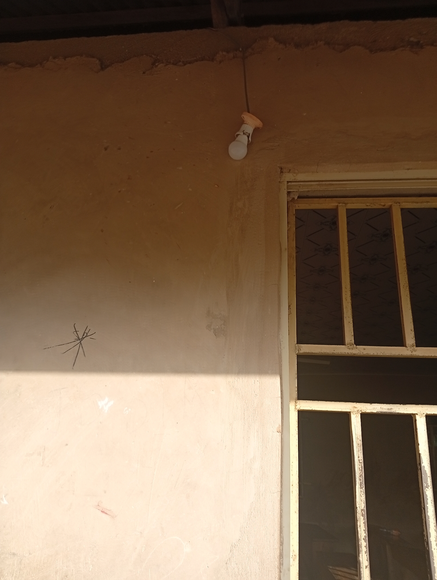 The image depicts an outdoor scene, likely in Jalingo, Nigeria. The primary subjects are a wall with a light fixture and a window. The wall is a rough, light-colored stucco material, with some darker marks and what appears to be a scratch or drawing of a star-like shape. A bare light bulb hangs from a wire near the top of the wall. To the right of the wall is a window with a metal security grill. The grill is painted a light yellow color and has vertical bars. The window itself appears to be dark, possibly covered by a screen or with the interior dark.
The lighting suggests it is daytime, with a strong shadow cast on the wall from an unseen object to the left, indicating sunlight. The overall impression is of a somewhat rustic or unfinished building. There are no people visible in the image. The scene is static and does not capture any specific activity or event. The location cues, such as the stucco wall and the style of the window grill, are consistent with what might be found in a residential or commercial building in Nigeria. There is no visible text in the image.