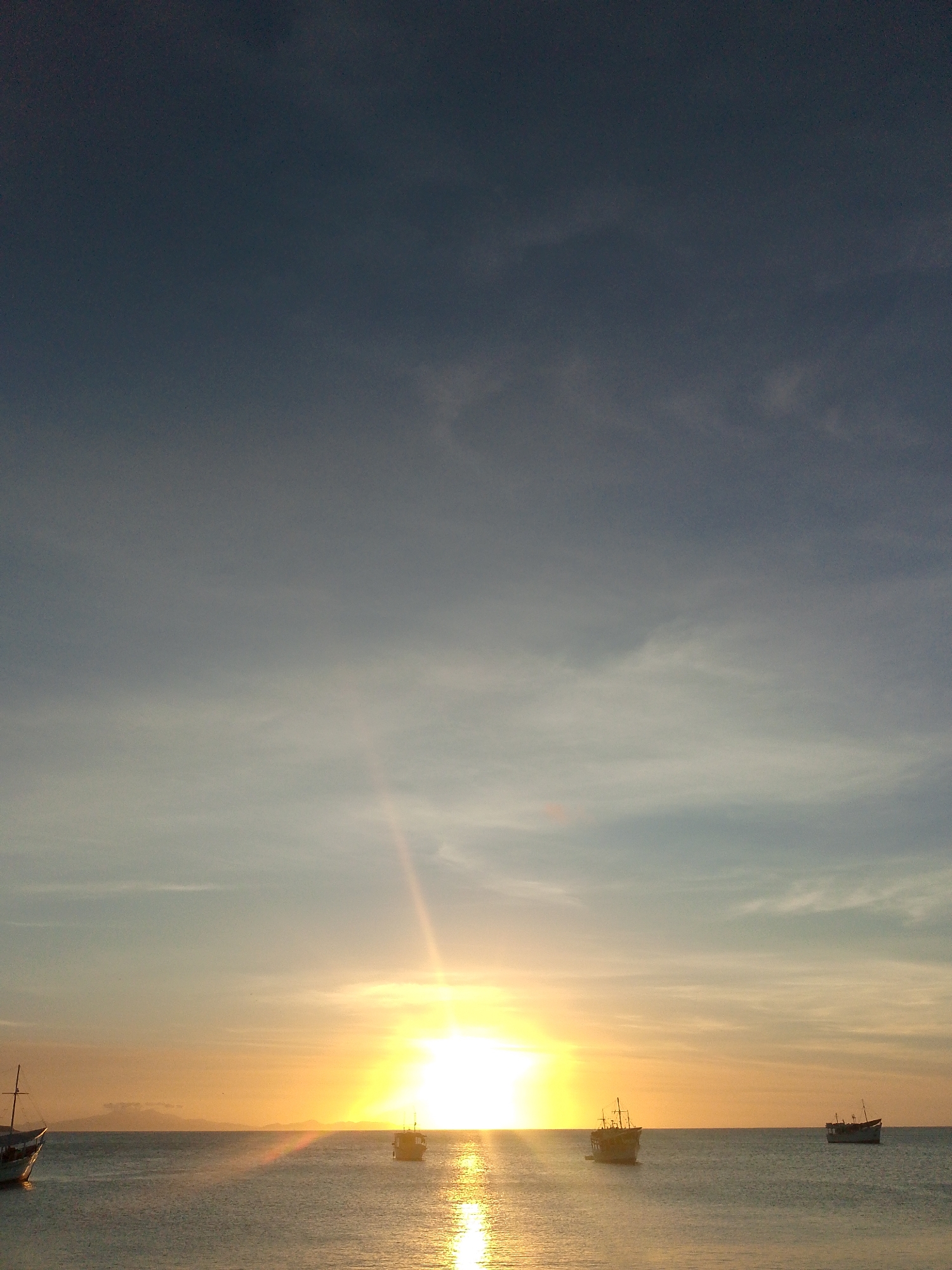 The image displays a panoramic sunset scene over the water in Juan Griego, Venezuela. The sky occupies the upper two-thirds of the frame, transitioning from a dark slate-grey at the apex to a lighter blue-grey mid-frame, with faint wisps of cirrus clouds discernible. The sun is positioned low and centrally on the horizon, emitting an intense, blinding golden-orange glare that stretches horizontally across the sky and creates a prominent, shimmering reflection path on the water's surface.
The body of water, likely the Caribbean Sea, occupies the lower third of the image. Its surface appears calm and displays a silvery-grey hue, reflecting the sky and the bright solar glare. Five distinct vessels are present on the water, rendered mostly in dark silhouette against the bright horizon and sun. From left to right: a boat with a visible mast is partially cropped on the far left; a second dark-hulled boat with a mast is positioned towards the left-center; a third, smaller dark-hulled vessel is situated near the center; a fourth dark-hulled boat with a mast is located in the right-center; and a fifth dark-hulled boat is visible on the far right. Distant landmasses or islands are faintly discernible as dark, hazy silhouettes along the horizon line, partially obscured by the sun's brilliance. No individuals or specific actions are visible within the frame.