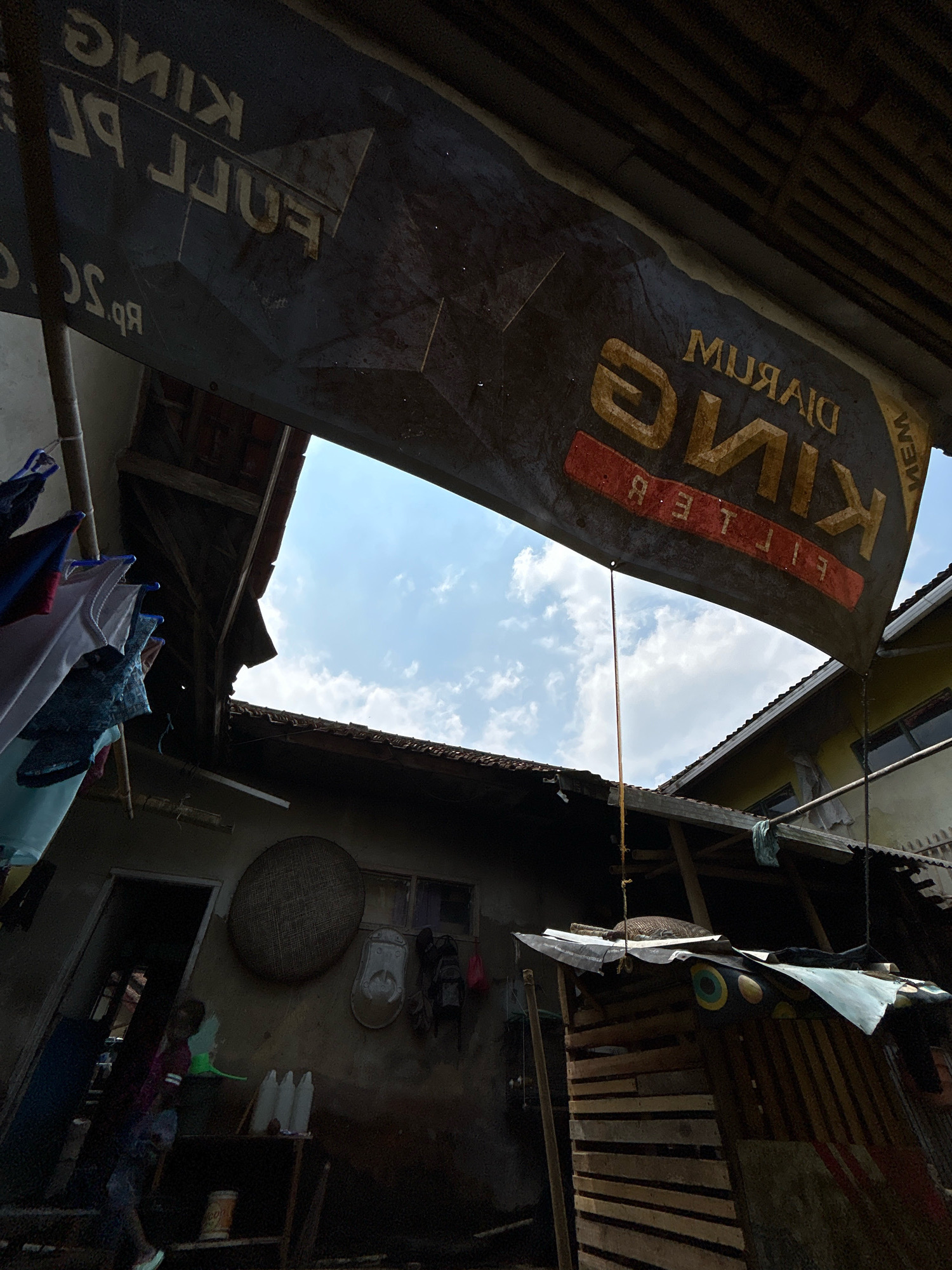 The image depicts an outdoor scene in a densely packed residential area, likely in Cihaurbeuti, Indonesia. A large, weathered banner displaying the words "DJARUM KING FILTER" is prominently hung overhead, casting a shadow over parts of the scene. The banner is torn and faded, suggesting prolonged exposure to the elements.
Below the banner, a glimpse of the sky is visible through an opening in the roof structure, revealing scattered white clouds against a blue sky. The surrounding structures appear to be makeshift, constructed from corrugated metal and wood. Laundry hangs on a line to the left of the frame, adding a domestic element to the otherwise utilitarian environment.
The ground level reveals a dark, cluttered area. A doorway leads into a dimly lit interior. A person, possibly a child, is partially visible in the doorway, wearing a pink and dark-colored garment. To the right of the doorway, a collection of white plastic bottles sits on a wooden shelf. A woven round object is mounted on the wall, and various items are hanging from it. A small, crudely built wooden structure with a sloped, covered roof stands to the right, adorned with a patterned cloth or tarp. The overall impression is one of a lived-in, modest dwelling in an urban or semi-urban Indonesian setting.