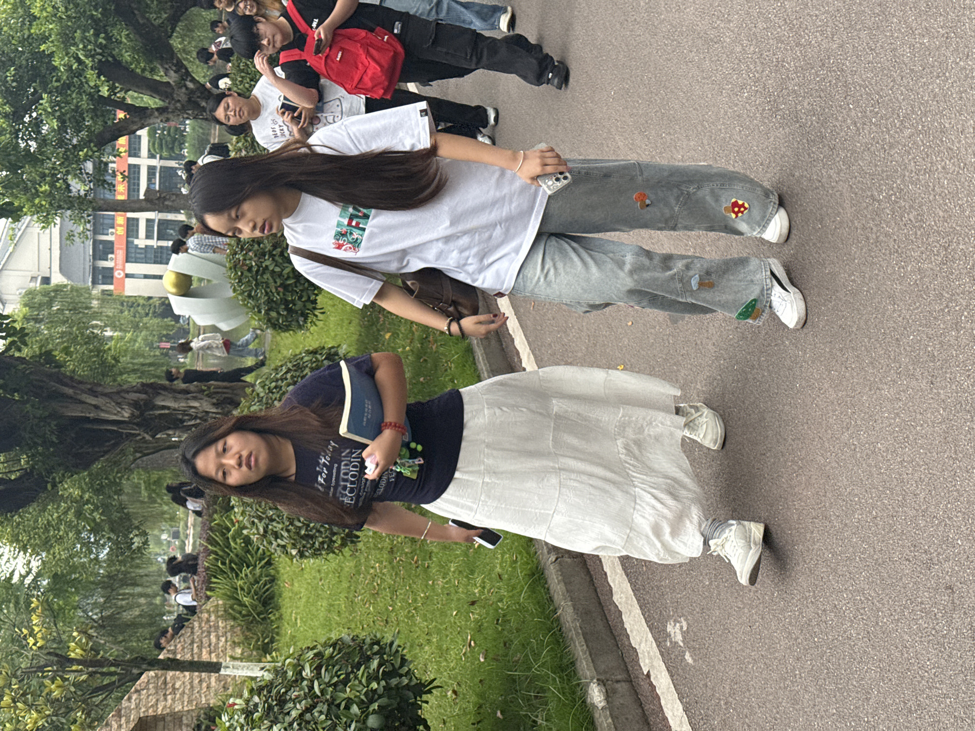 Two women are walking on a pathway. One wears a white t-shirt, blue jeans with mushroom patches, and white sneakers. The other wears a dark t-shirt, a long white skirt, and white sneakers, and is holding a book. They are on a paved path next to a grassy area with trees. Other people are visible in the background, as well as some buildings. The location is Jiangjin, China.