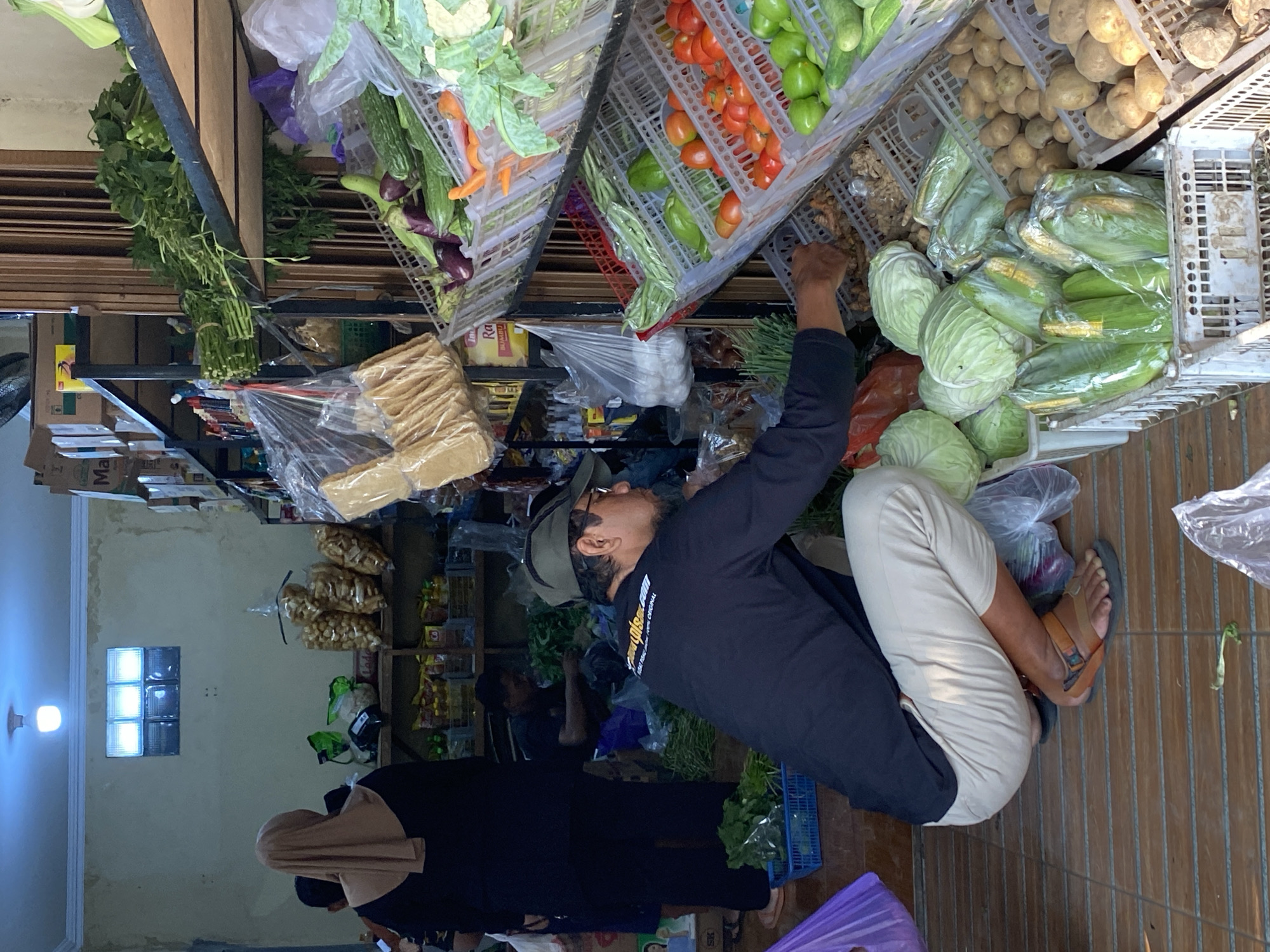 A man wearing a dark long-sleeved shirt, beige pants, a green cap, and sandals is crouching in a market stall in Kasihan, Indonesia. He appears to be sorting through produce, reaching into a crate of brown, knobby items. In front of him are several plastic-wrapped ears of corn and cabbage heads. To his right, shelves are laden with various vegetables in plastic crates, including tomatoes, cucumbers, eggplants, and what appear to be chilies. Behind him, a woman wearing a tan hijab and a dark outfit stands near shelves stocked with bagged snacks. Another man in a dark shirt is visible in the background, also tending to produce. The stall features wooden shelving units and tiled flooring. Lighting appears to be a mix of overhead fluorescent and natural light filtering through a small window.