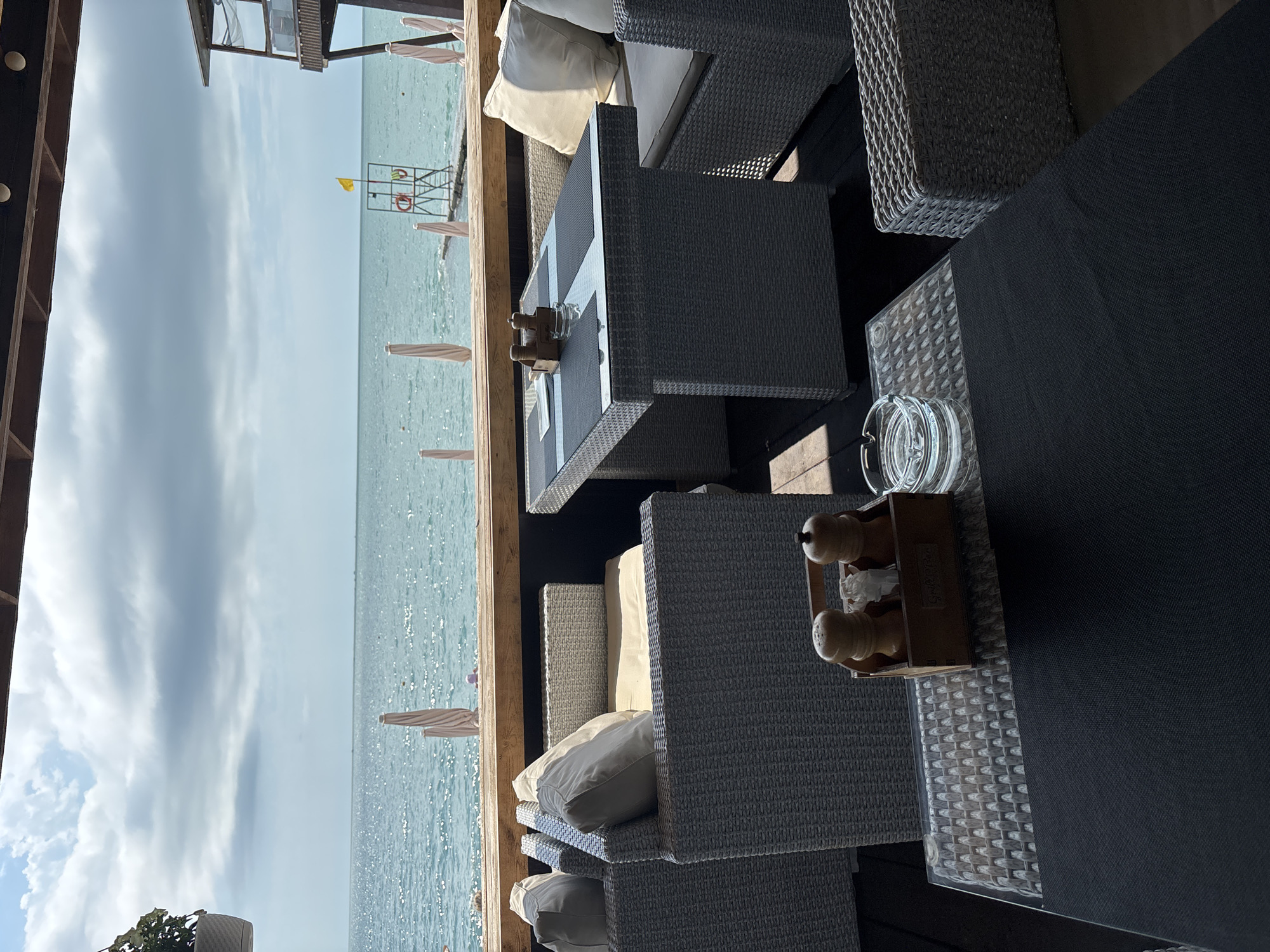 This image depicts an outdoor seating area of a restaurant or cafe with a view of the sea. The perspective is from inside the seating area, looking out towards the water. The scene is set on a wooden deck, furnished with several dark grey wicker chairs and tables, some with glass tops. Cushions in neutral tones are placed on the chairs. One table has an ashtray and a wooden condiment caddy containing salt and pepper grinders.
In the background, the sea is visible, with a light blue sky above, suggesting a daytime scene, likely morning or afternoon. The water appears calm with some light rippling. Along the shoreline, several closed white umbrellas are lined up. Further out, a lifeguard station with a yellow flag and life preservers can be seen. There are no people clearly visible in the immediate seating area. The overall atmosphere suggests a relaxed and pleasant environment for dining or lounging by the sea.
The image is oriented vertically, and the lighting indicates the sun is casting shadows, suggesting a sunny day with some clouds in the sky. The setting appears to be a coastal resort or a beachfront establishment, possibly in Sochi, Russia, given the context. No discernible text is visible in the image.