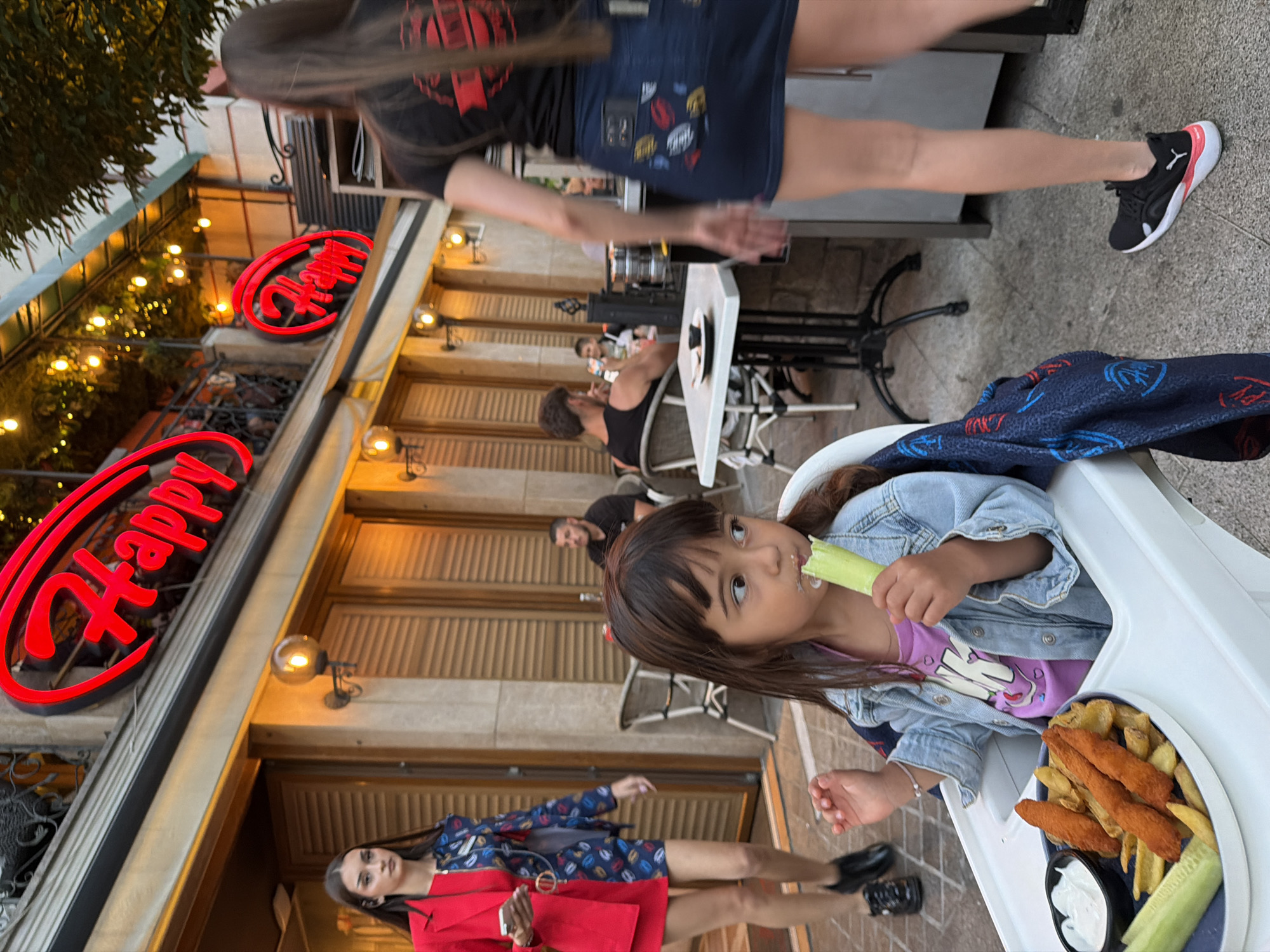 In Burgas, Bulgaria, outside a restaurant with red neon "Happy" signs, a young girl sits in a high chair, eating a celery stick and french fries with a bowl of dip. A woman in a red jacket walks past in the background. Another woman with long hair is walking away from the camera. Other patrons are seated at tables. The scene is outdoors, with tables and chairs set up on a patio.