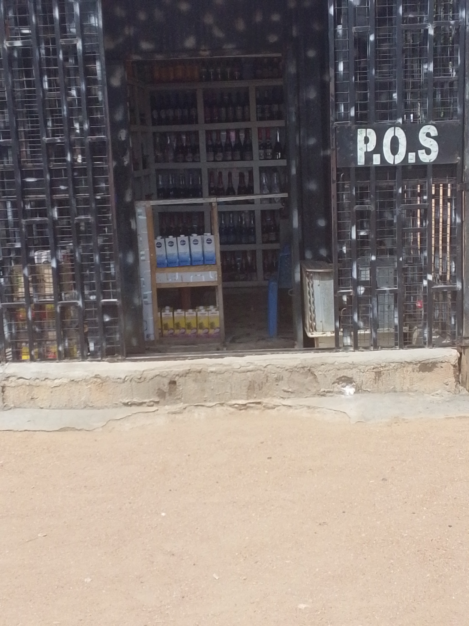 The image shows the exterior of a small shop in Jalingo, Nigeria. The shop has a metal grate front and a sign that reads "P.O.S". Inside the shop, bottles of beverages are displayed on shelves. A blue plastic chair is visible in the background. The ground in front of the shop is dusty and sandy. The overall scene suggests a typical street-level business in a Nigerian city.