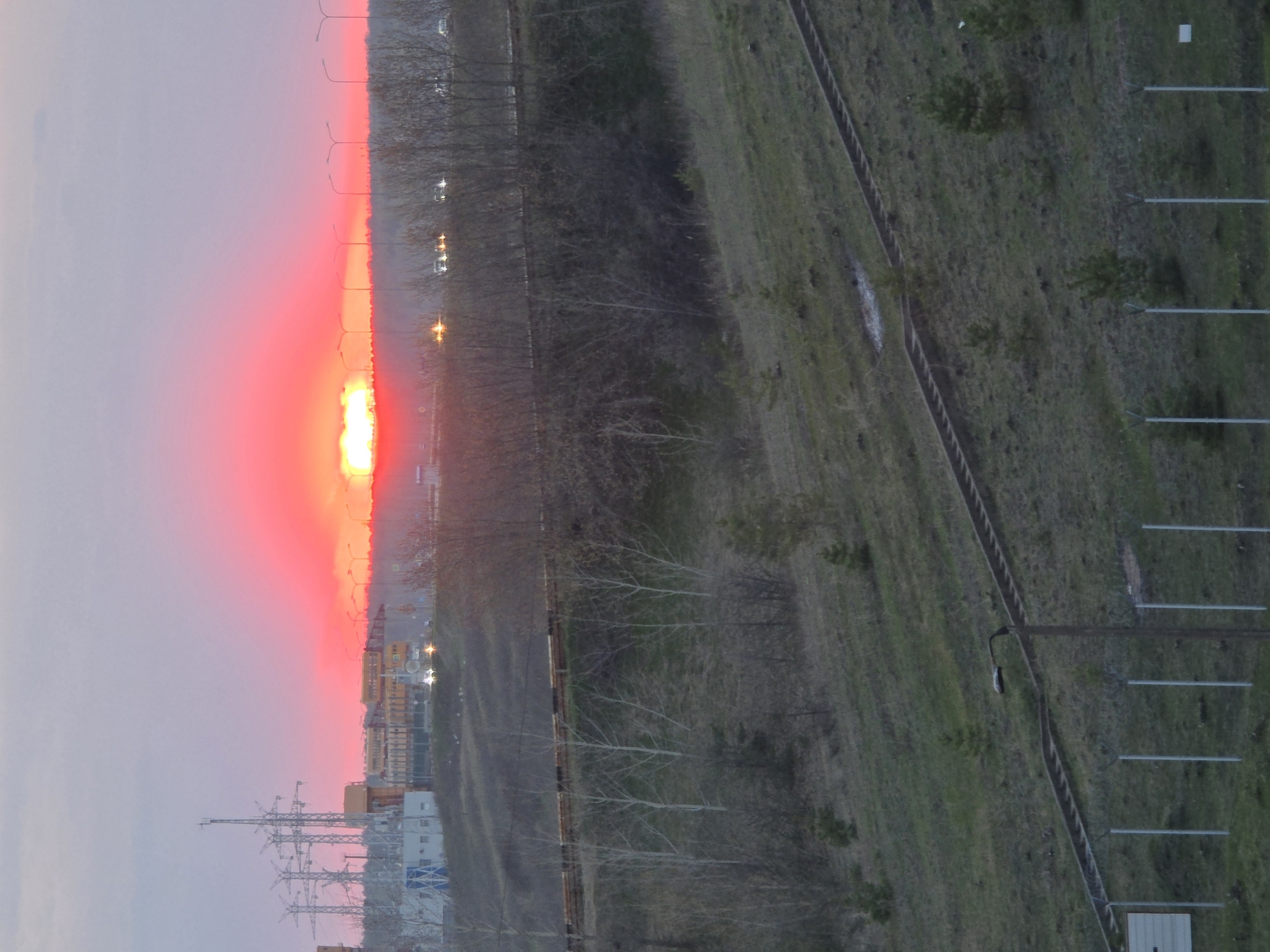 The image shows an aerial view of a landscape in Chaikovsky, Russia, at sunset. The sky is a blend of gray and vibrant red-orange where the sun is setting, casting a glow over the horizon. In the foreground, there are power lines and some buildings. A road with streetlights lines the edge of the city area, separating it from a green, grassy area on the right. A fence and some light poles run along the grassy area.