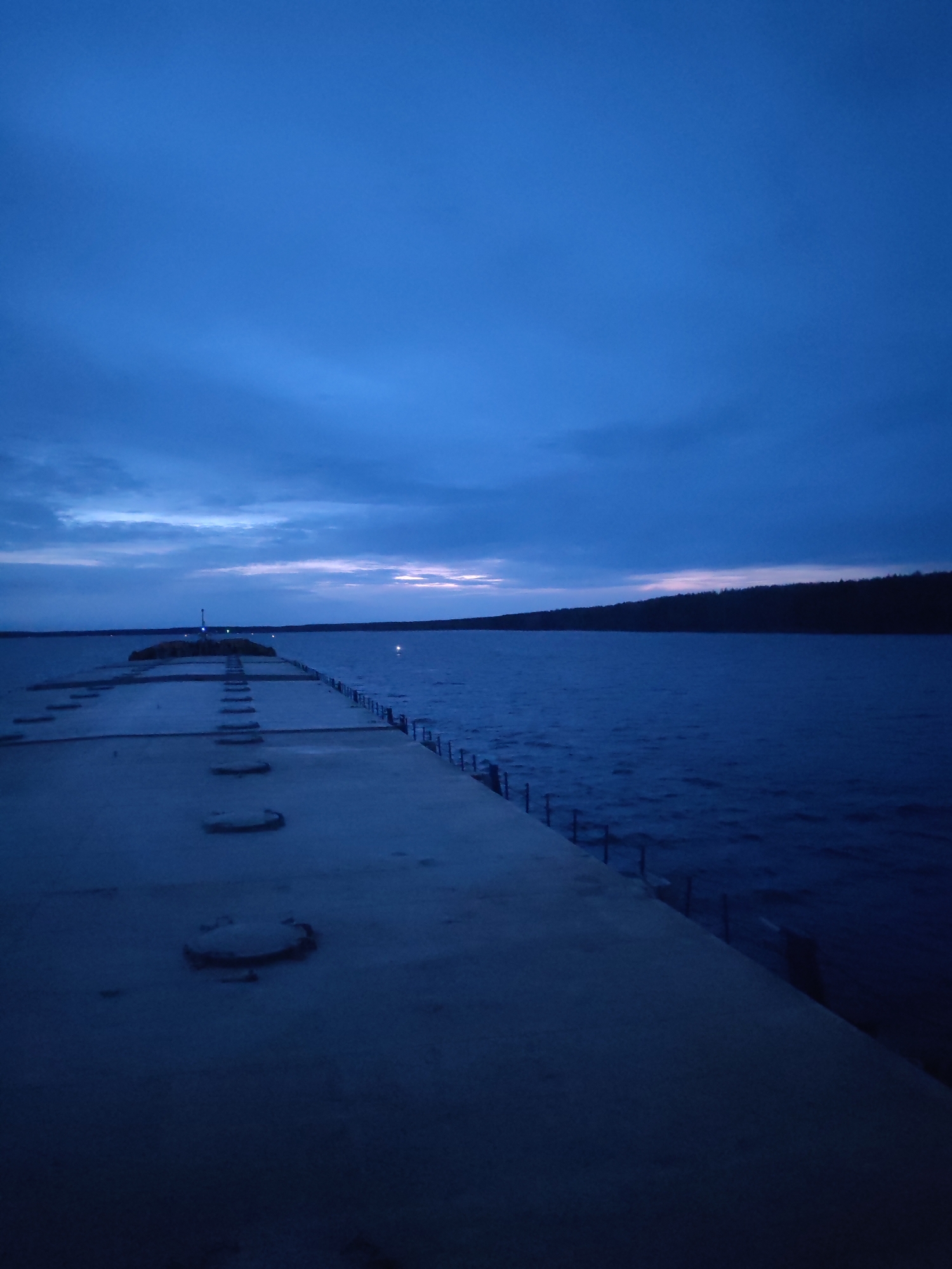 A low-angle, long-exposure photograph captures a concrete pier extending into a body of water, likely taken at twilight or dawn. The pier is the primary subject, and it stretches out from the foreground towards a distant structure with a light source, possibly a lighthouse or marker. The pier has several circular indentations along its surface. A metal railing lines the side of the pier closest to the water. The water appears choppy with small waves. On the opposite side of the water is a dark shoreline of trees. The sky is mostly covered in dark blue clouds. This image appears to be taken in Topornya, Russia.