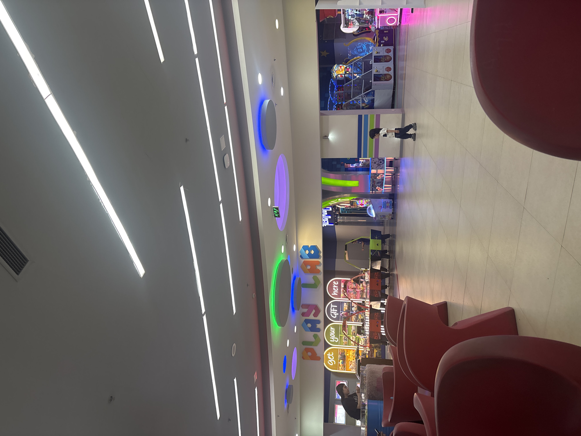 The image captures an indoor amusement or arcade center, likely within a mall, with bright artificial lighting and a clean, modern aesthetic. The scene is dominated by a variety of arcade games on the right side and a spacious, tiled walkway.
Several subjects are visible. A small child with dark hair, wearing a light-colored top and dark trousers, walks across the light-colored tiled floor, moving towards the arcade machines. In the foreground, on the far right, parts of several red, modern-shaped chairs or seating units are visible. An adult with dark hair and a dark top is seated near these chairs, partially obscured.
The setting is characterized by rows of arcade machines featuring flashing lights and colorful graphics, including claw machines and basketball-themed games. Above these games, a large, colorful sign spells out "PLAYLAB" in stylized, multi-colored letters. Below this, a smaller sign reads, "Get your GIFT here." Another green illuminated sign on the ceiling indicates "EXIT."
The ceiling is a notable feature, with numerous long, linear fluorescent lights providing bright illumination. Additionally, there are several circular recessed lights with vibrant, color-changing LED rings in shades of blue, purple, and green, adding to the playful atmosphere. An air vent is also visible on the ceiling. The floor is covered in light-toned square tiles. The overall impression is that of a lively, family-friendly entertainment venue located in Yerevan, Armenia.