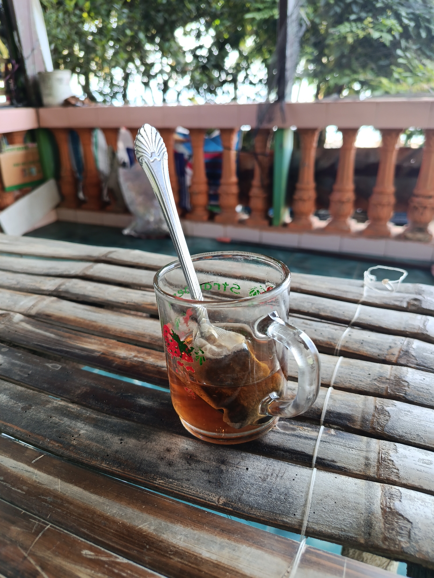 A clear glass mug filled with amber-colored tea and a tea bag rests on a weathered bamboo table. A silver spoon with a decorative, shell-like handle is placed inside the mug, partially submerged in the tea. The mug has a delicate floral pattern with red and green accents around its rim. In the background, a wooden railing with vertical balusters is visible, beyond which lush green foliage and the blurry shapes of trees can be seen, suggesting an outdoor setting, possibly a balcony or veranda. The overall lighting suggests daytime. The image appears to have been taken in Mansalean, Indonesia, a location known for its tropical climate and natural beauty, consistent with the verdant foliage. There are no people present in the image.