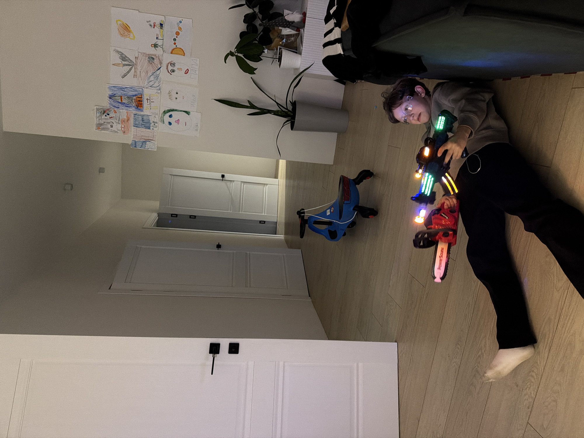 The image depicts a domestic indoor scene in Chaikovsky, Russia, likely a living room or hallway. A young boy, wearing glasses, a grey sweatshirt, dark pants, and a white sock, is lying on a light wooden floor. He is actively engaged in play, holding two illuminated toy weapons: a large, futuristic blaster with prominent green, blue, and yellow LED lights, and a smaller red toy chainsaw, which also glows and has "Power Sound" printed on its side. His expression is focused or slightly curious as he looks towards the viewer.
Nearby, a blue ride-on toy, resembling a tricycle or scooter hybrid, rests on the floor. The background features light-colored walls and several white interior doors, some with black handles. On one wall, a collection of colorful children's drawings is displayed, including various portraits and other illustrations. Visible Russian text on some drawings includes "ПАПА" (Papa/Dad) and "ИЛЬЯ" (Ilya), with another word possibly being "МАМА" (Mama/Mom). Potted plants are positioned on the floor and a shelf against the wall. The lighting suggests an indoor setting, possibly in the evening or a dimly lit space, illuminated by artificial light sources.