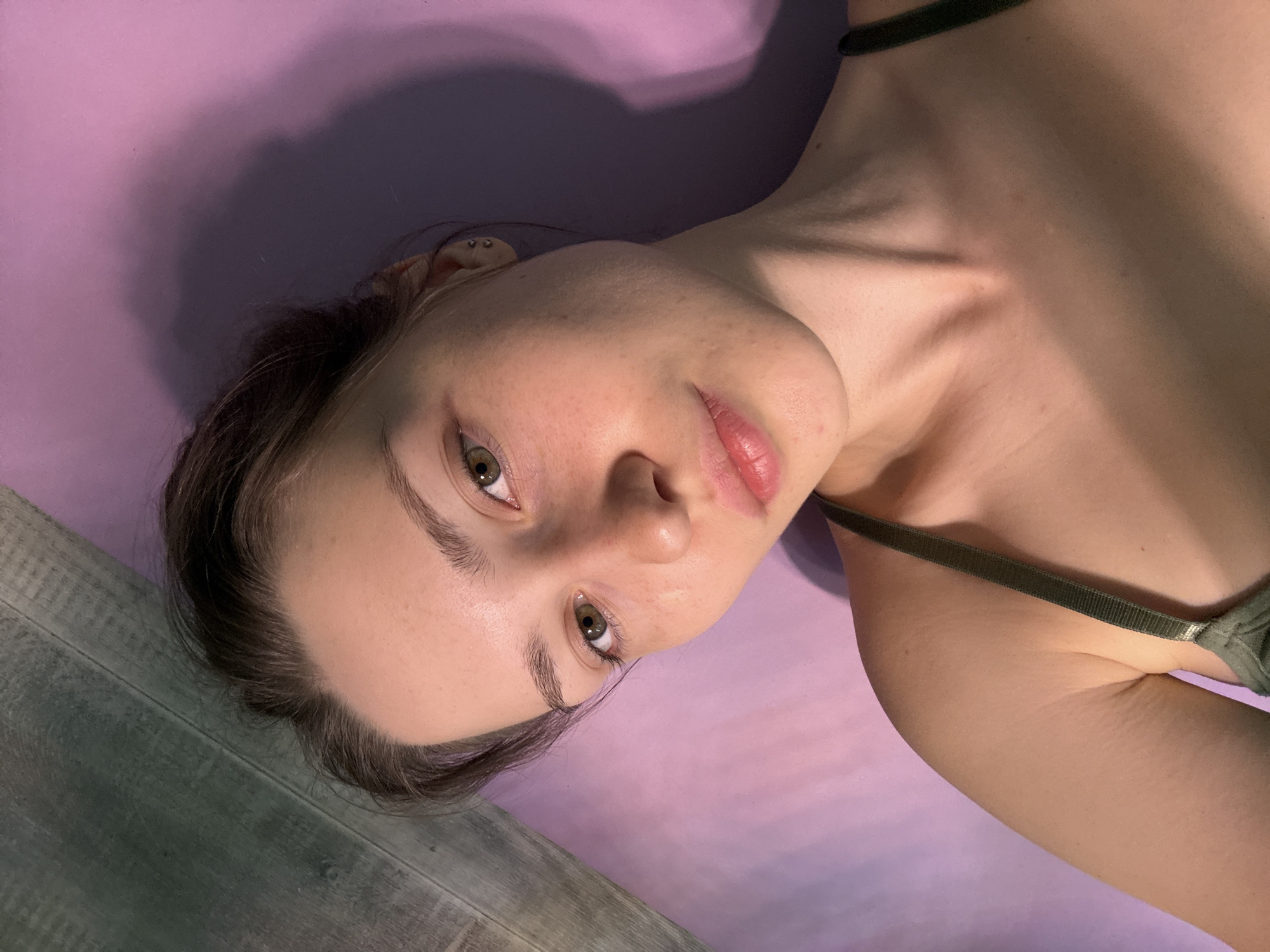 An overhead, slightly tilted close-up portrait shows a young individual, likely female, lying on their back and looking directly into the camera. The subject has fair skin with visible pores and minor surface irregularities. Her eyes are light brown or hazel, framed by dark, well-defined eyebrows. Her lips are slightly parted, exhibiting a natural pink hue. Dark brown hair is pulled back, with finer strands visible around the forehead and temples. A small hoop earring is visible in the lobe of her left ear, accompanied by what appears to be a small stud piercing higher on the helix.
The subject is wearing a dark olive-green strap top or bra, with the strap visible over her right shoulder. Her prominent collarbones and a portion of her upper arm are also visible in the frame. She is positioned on a muted purple or lilac-colored surface, consistent with a yoga mat or similar padded surface. A soft, dark shadow is cast across this purple surface, primarily behind the subject's head (relative to the image's orientation), suggesting an overhead light source. To the lower left of the frame, a section of dark greyish-green wooden plank flooring is visible, indicating the mat is placed on a floor. The lighting is soft and even, illuminating the subject's face and upper body. The scene is located indoors, in the city of Saint Petersburg, Russia.