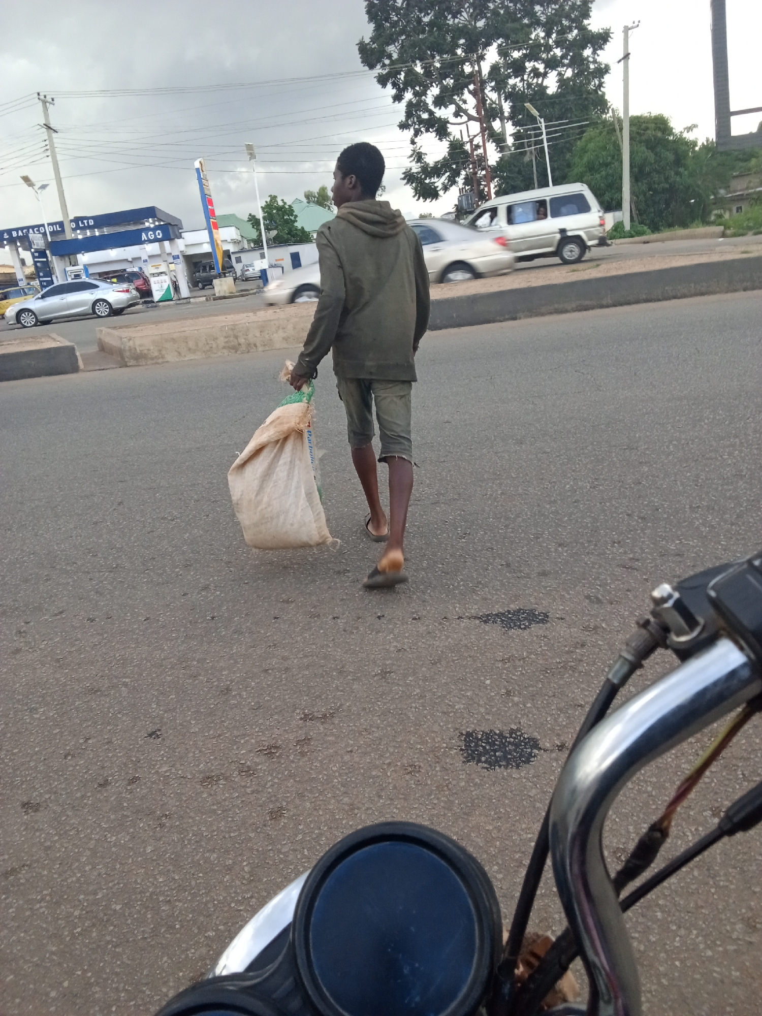 A young Black male, wearing a faded green hooded sweatshirt and olive-green shorts, walks on an asphalt road in Jos, Nigeria, carrying a large, light-colored sack in his right hand. He is walking away from the camera, towards a gas station with blue and white branding that reads "BABACO OIL LTD" and "AGO". Several cars are visible at the gas station and on the road, with one silver sedan and a white van in motion. The sky is overcast, and the surrounding environment includes trees and power lines. The perspective suggests the photo was taken from a moving vehicle, possibly a motorcycle, as part of its handlebars and a mirror are visible in the foreground.