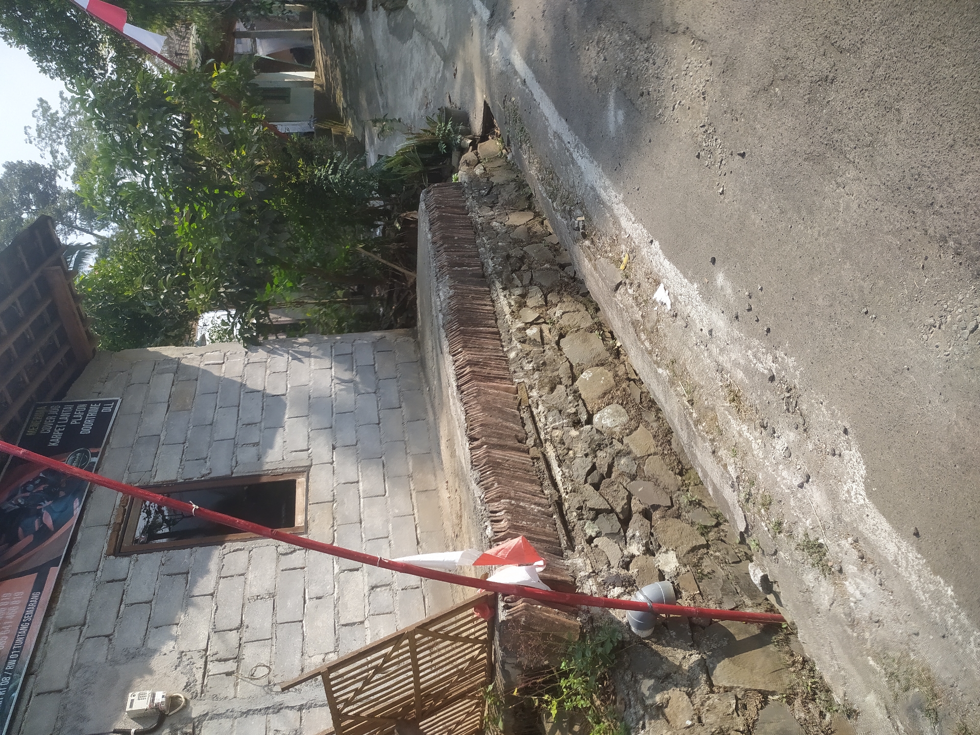 The image shows a street in Lopait, Indonesia. A red pole with a red and white Indonesian flag attached to it stands in front of a cinderblock building. The building has a small window and a meter on the exterior wall. A drainage ditch runs alongside the street, filled with rocks and some vegetation. The street is paved and appears to be in a rural or suburban area, with trees and other buildings visible in the background. The sun is shining, casting shadows on the scene.