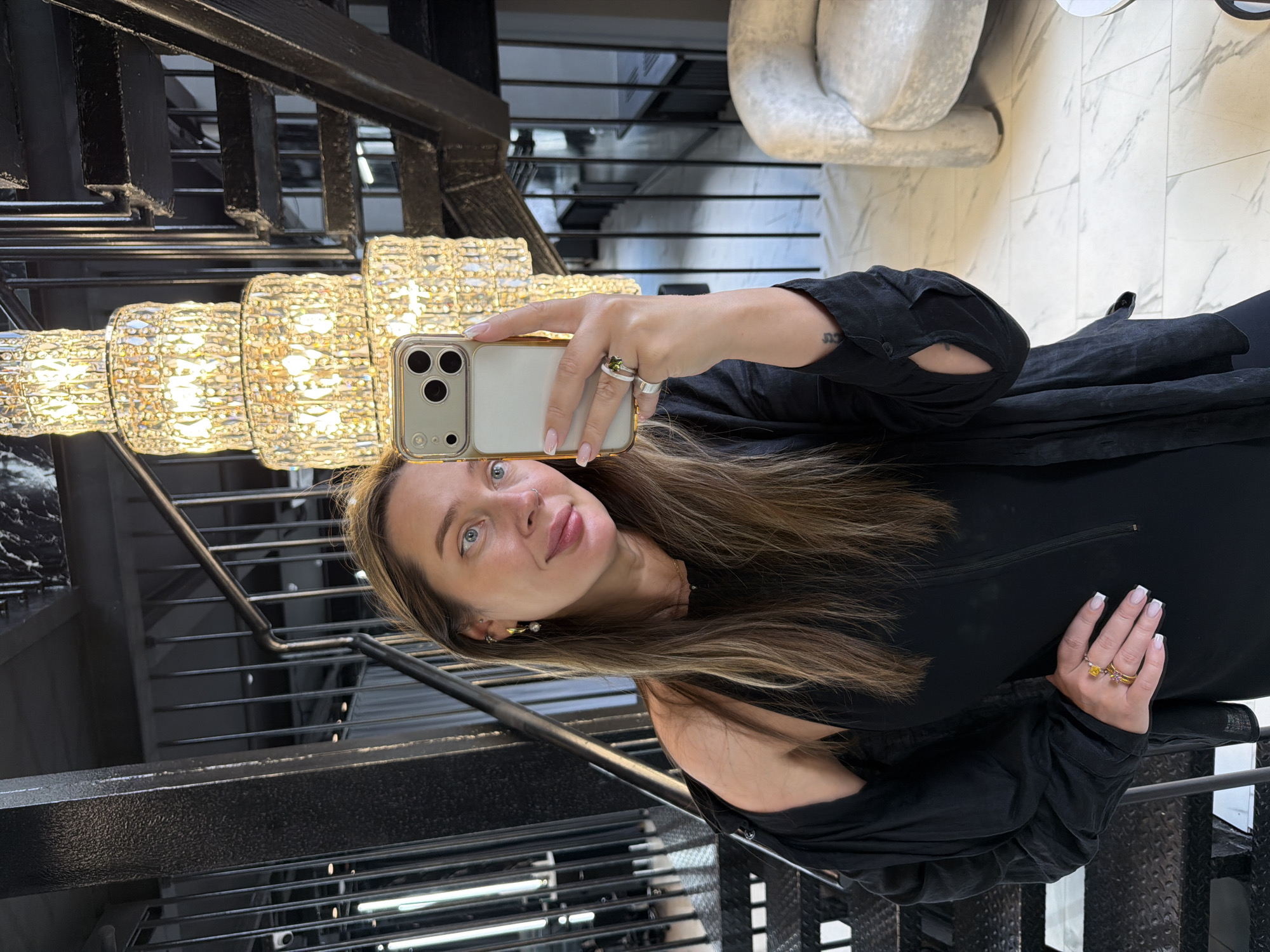 A woman with long, highlighted brown hair is taking a selfie while standing on a black metal staircase with a large, multi-tiered chandelier. She is wearing a black outfit and has a phone with a gold case in her hand. Her other hand, also adorned with rings, is resting near her waist. The background includes a glimpse of a white, marble-like floor and a white, rounded piece of furniture. The location is unspecified except for Podolsk, Russia.