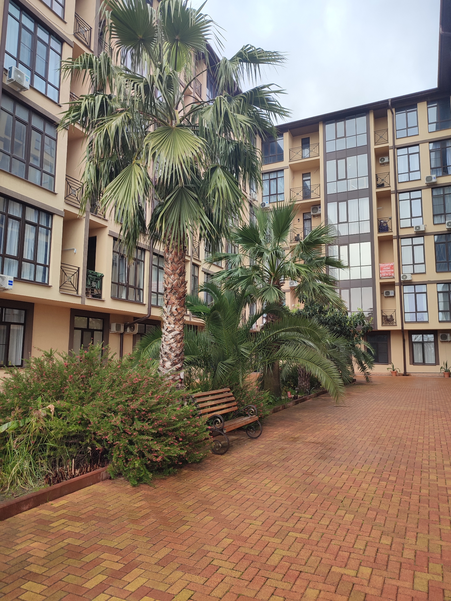 The image depicts an outdoor courtyard in what appears to be a residential complex. The location is identified as Sochi, Russia, due to the architectural style and vegetation.
The courtyard is paved with red and yellow patterned bricks. Several palm trees are present, interspersed with bushes and other plants. A wooden bench with ornate metal legs sits near the shrubbery on the left side of the frame.
Two beige-colored buildings surround the courtyard. Both buildings have dark-framed windows and small balconies with dark metal railings. Air conditioning units are visible on the exterior walls of both buildings. A small sign is visible on one of the buildings.
The sky is overcast. There is no presence of people.