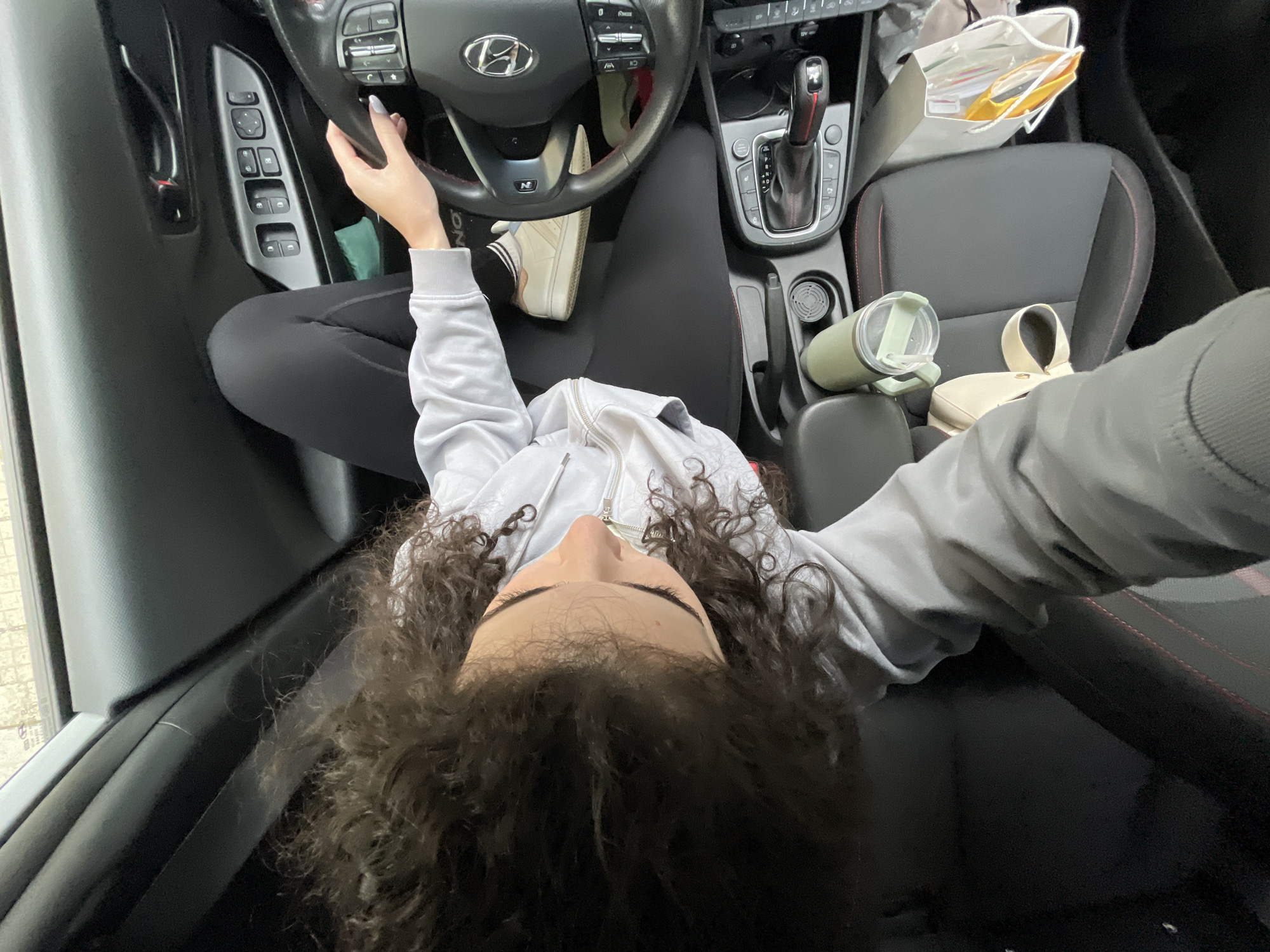 A woman with curly brown hair is seated in the driver's seat of a Hyundai car, possibly a Hyundai i30 N based on the steering wheel. She is wearing black leggings and a light gray zip-up hoodie. Her left hand is on the steering wheel, and there is a white sneaker on the floor near her left foot. A green water bottle and a beige purse are on the center console, beside the passenger seat where there is a white shopping bag. The location is a car interior, but Sofia, Bulgaria, cannot be confirmed from the image.