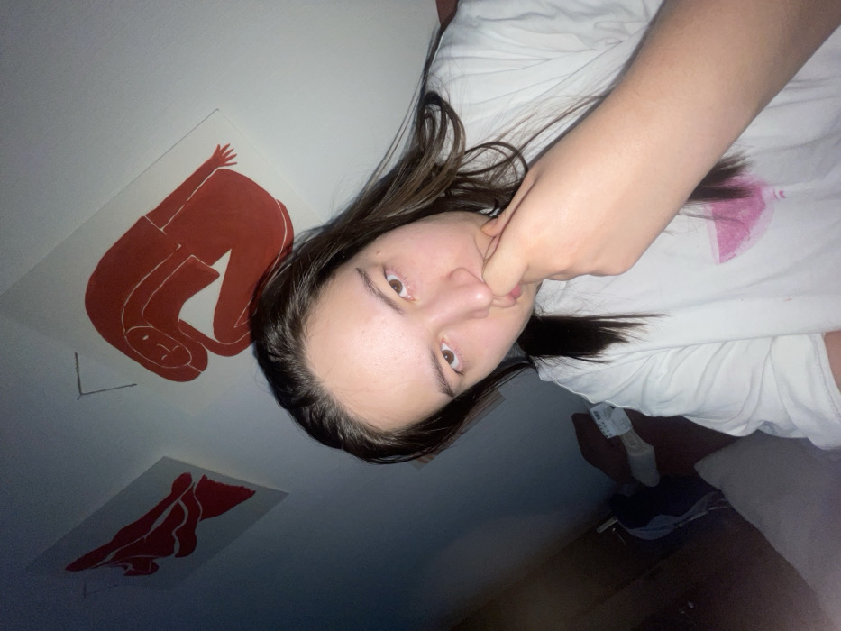 A woman with long dark hair, wearing a white shirt with a pink stain, looks at the camera while putting her fist near her mouth. On the wall behind her are two red prints of human figures. The room appears dimly lit. In the background, partial views of clothing and what appears to be a bed are visible.