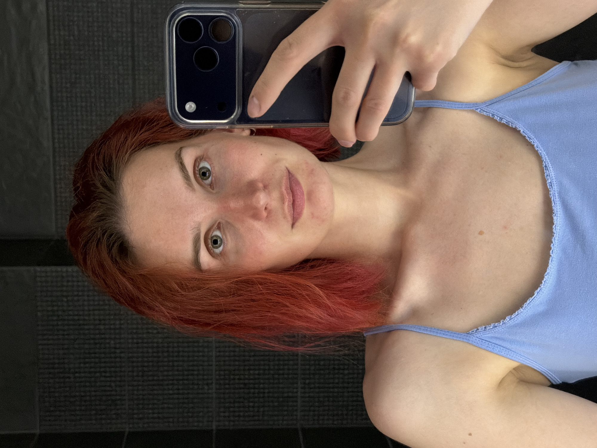 A woman with red hair takes a selfie in a bathroom with dark grey tiled walls. She is wearing a light blue tank top. The phone she is using is a dark color with a clear case. The location is unknown, and there is no indication that it is in Chaikovsky, Russia.