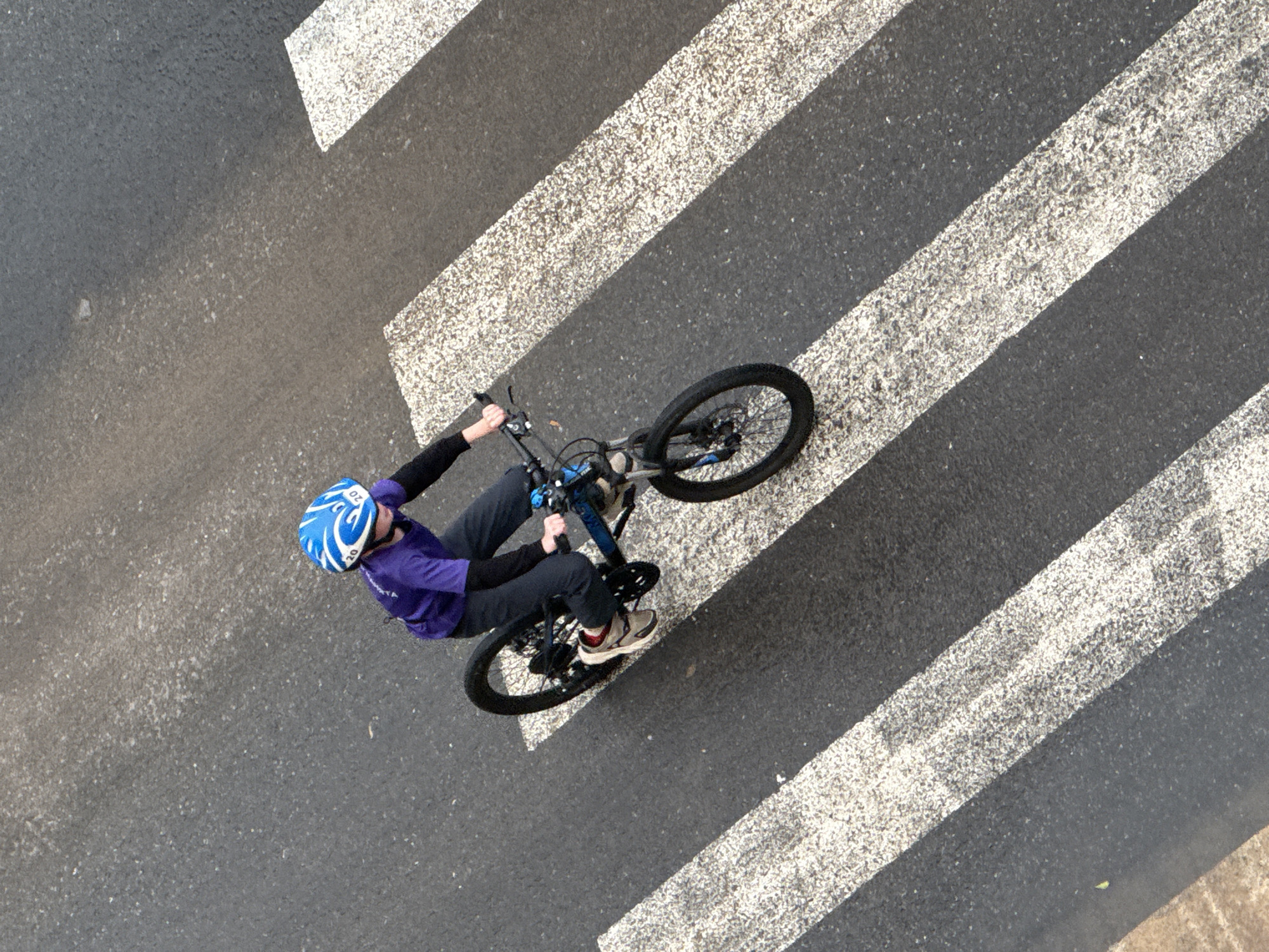 An overhead, high-angle shot captures a young person wearing a blue helmet and a purple t-shirt, riding a bicycle across a pedestrian crossing. The bicycle is dark-colored with silver accents. The pedestrian crossing consists of parallel white stripes on a dark asphalt road. The person is positioned on the right side of the bicycle, with their left foot on the pedal. The scene appears to be an urban environment in Votkinsk, Russia.