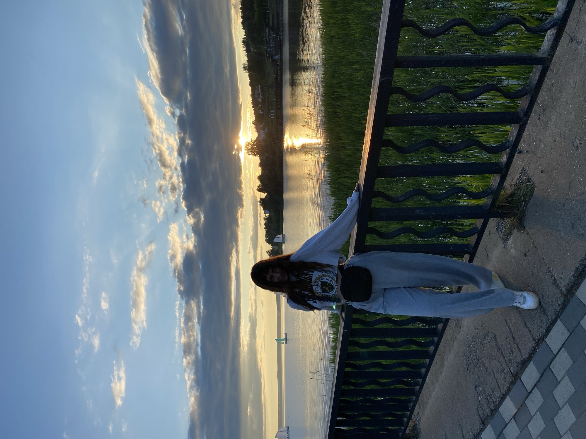 The image depicts a young woman posing on a waterfront promenade during sunset. She stands leaning casually against a dark metal railing with distinctive wavy top bars, looking towards the camera with a slight smile. She has long, dark hair with bangs and is dressed in a light grey long-sleeved sweatshirt, matching grey sweatpants, and white sneakers. A black crossbody bag is worn across her chest. On her sweatshirt, the text "CALIFORNIA STATE" is visible as part of a graphic design, with other words partially obscured below it.
Behind the woman and the railing, a wide body of water, likely a lake or wide river, stretches towards the horizon. The setting sun is low on the left, casting a brilliant golden reflection across the water, creating a shimmering path of light. Tall green reeds or grass are visible in the water near the foreground bank. On the distant shore, a line of trees and scattered buildings are silhouetted against the bright sky. The sky above is clear blue, transitioning to warmer tones near the horizon, with fluffy white and grey clouds scattered across it, illuminated by the low sun. The foreground shows a concrete walkway and a patterned tiled path. The overall scene suggests a calm, pleasant late afternoon or early evening by the water.