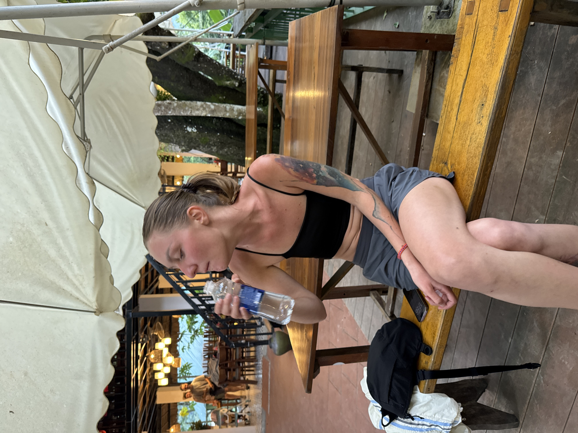 A young woman with her hair pulled back in a ponytail is sitting on a wooden bench, drinking from a clear plastic water bottle. She is wearing a black crop top and grey shorts, and has a tattoo on her left arm. A black backpack and a white garment are on the bench next to her, as well as a mobile phone. The bench is on a wooden deck, with more wooden tables and chairs visible in the background. Behind the tables, there are trees and lush greenery. The scene appears to be an outdoor dining area or rest stop in a tropical or subtropical location, possibly Cổ Lũng Commune, Vietnam, given the presence of a traditional-looking vase and the overall ambiance. The lighting suggests it is late afternoon or early evening.