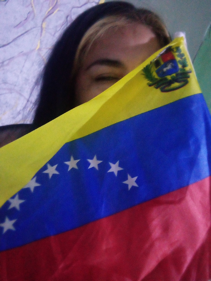 A woman partially hides her face behind a flag of Venezuela. The flag displays horizontal stripes of yellow, blue, and red with an arc of eight white stars on the blue stripe. The Venezuelan coat of arms is on the upper left of the yellow stripe. Only the woman’s eye and dark hair with a blonde streak are visible. The background is a light, textured wall.