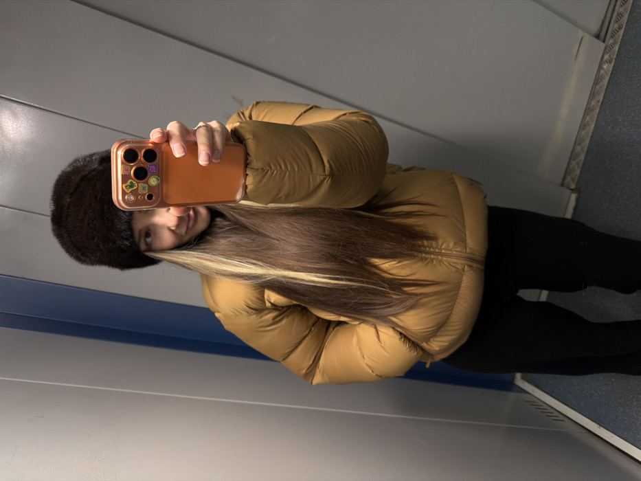 A female individual takes a self-portrait photograph in an enclosed, utilitarian interior space within Khimki, Russia. The environment features vertical grey wall panels, a narrow blue vertical strip on the left, and a dark blue floor with a metallic threshold, consistent with a public transport vehicle or elevator cabin. The subject is wearing a dark brown, possibly fur, winter hat, a mustard-yellow puffer jacket, and black pants. Her long, dark brown hair with blonde highlights extends over her shoulders. She holds a smartphone in her right hand, extended towards the viewer, enclosed in a translucent orange case that features distinct camera cutouts and multiple small, colorful decorative stickers. A ring is visible on her left ring finger. Her expression is a slight, casual smile.