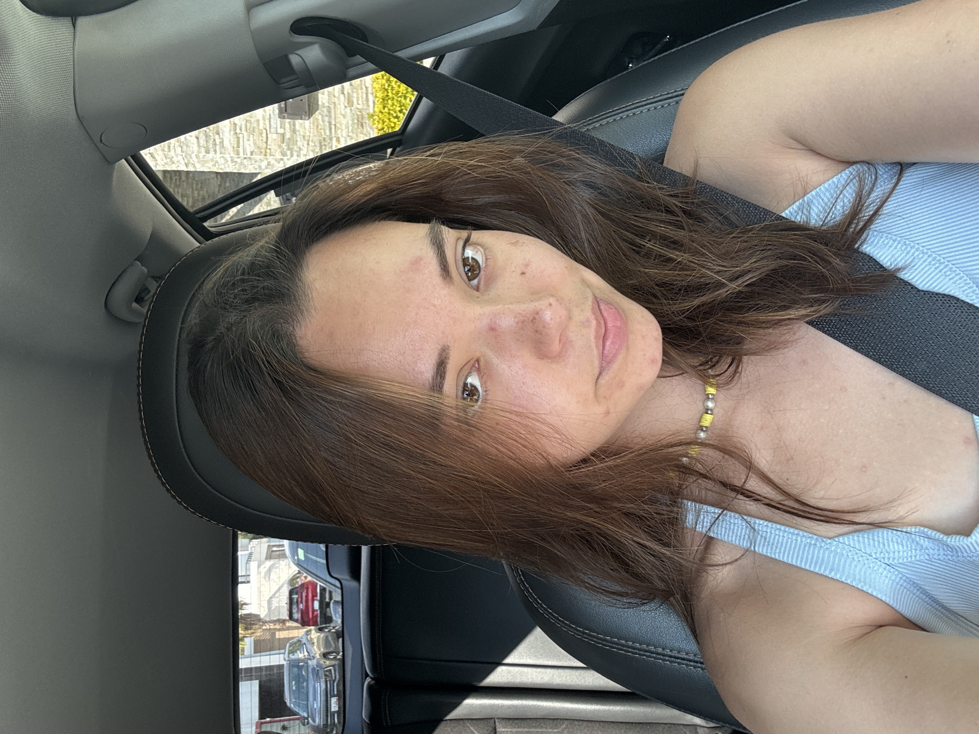 A woman is seated in the driver's seat of a car, wearing a seatbelt and a light blue tank top. She has brown hair and brown eyes and is facing the camera. The interior of the car is visible, including the dashboard and seats. The environment outside the car is visible through the windows, including buildings and other vehicles. The specific location in the city of San Bernardino Tlaxcalancingo, Mexico is not definitively discernible from the provided image.