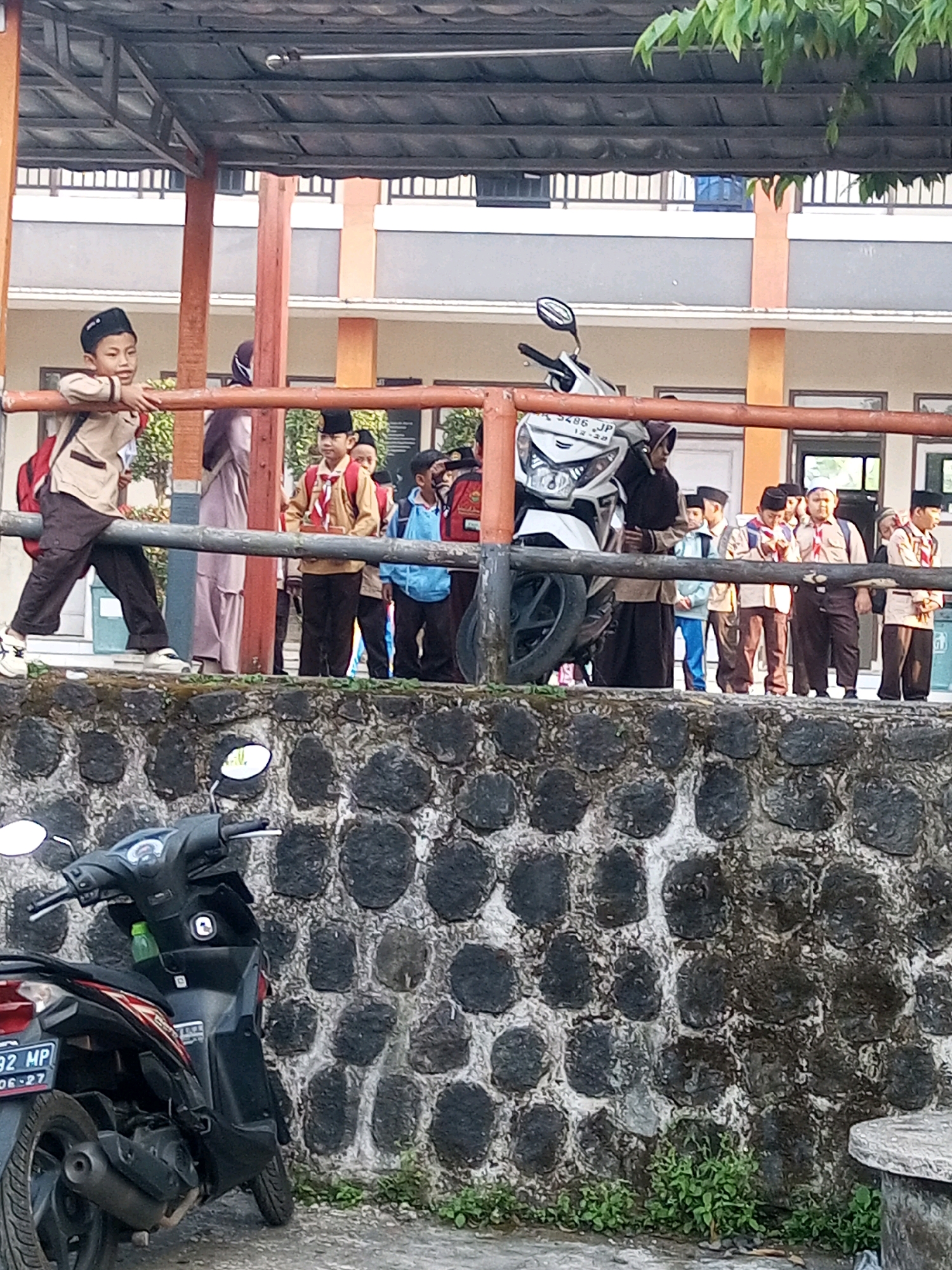 The image shows a group of uniformed schoolchildren and a white motorcycle in front of a building in Krasak, Indonesia. The children, wearing khaki uniforms and black caps, appear to be of elementary school age. One child in the foreground is leaning on a railing, looking towards the left. Others stand in a line, some holding small objects, possibly awards or flags. The building behind them has a light-colored facade with orange accents and a dark roof. A stone wall with vegetation at its base runs across the lower part of the image, with two black motorcycles parked in front of it. The scene suggests a school event or gathering.