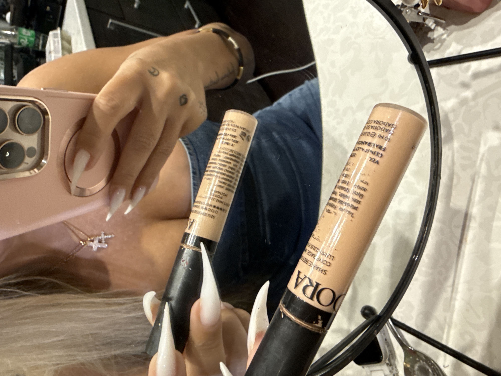 The image depicts a close-up, self-shot of a person holding two tubes of makeup. The person's hands, with long, white acrylic nails, are visible, as is a portion of their blonde hair, a necklace with a cross pendant, and a glimpse of a pink phone. The makeup tubes are positioned in the foreground, with one tube being held by a hand and the other resting in front of it. The tubes appear to be concealers or foundations, with text indicating a brand name starting with "DORA". The background shows a blurry kitchen environment with cabinets and a countertop. The scene is indoors, and there is no indication of the city of Odesa, Ukraine, or any specific geographical location. The lighting suggests an indoor setting, likely a home or a personal space, with artificial light sources. The focus is on the makeup products and the person's hands.