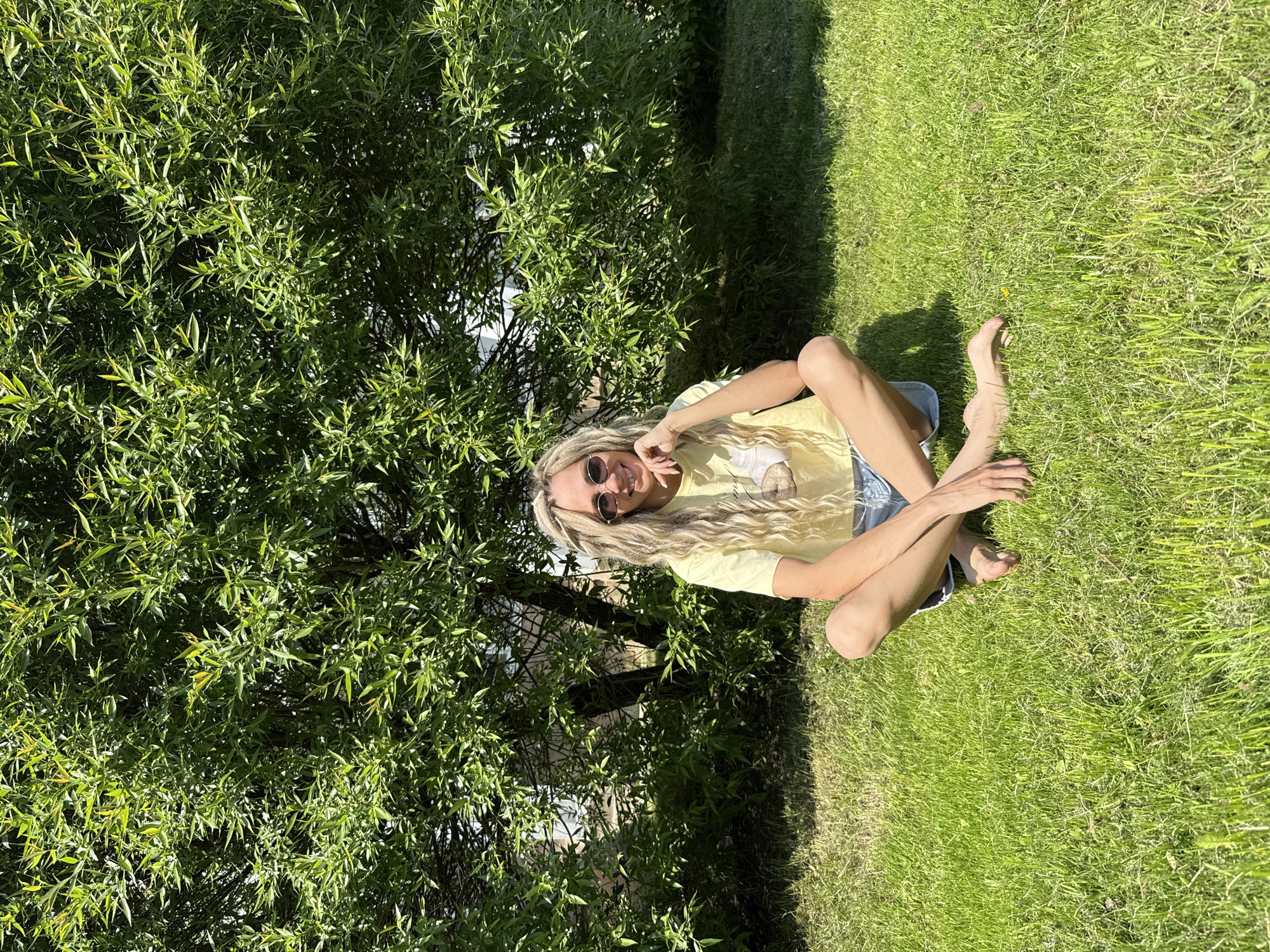 A blonde woman wearing sunglasses, a light yellow t-shirt, and denim shorts is sitting on green grass in an outdoor setting. She is positioned to the right of a large green bush or tree. The location is presumably Rusanovo, Russia.